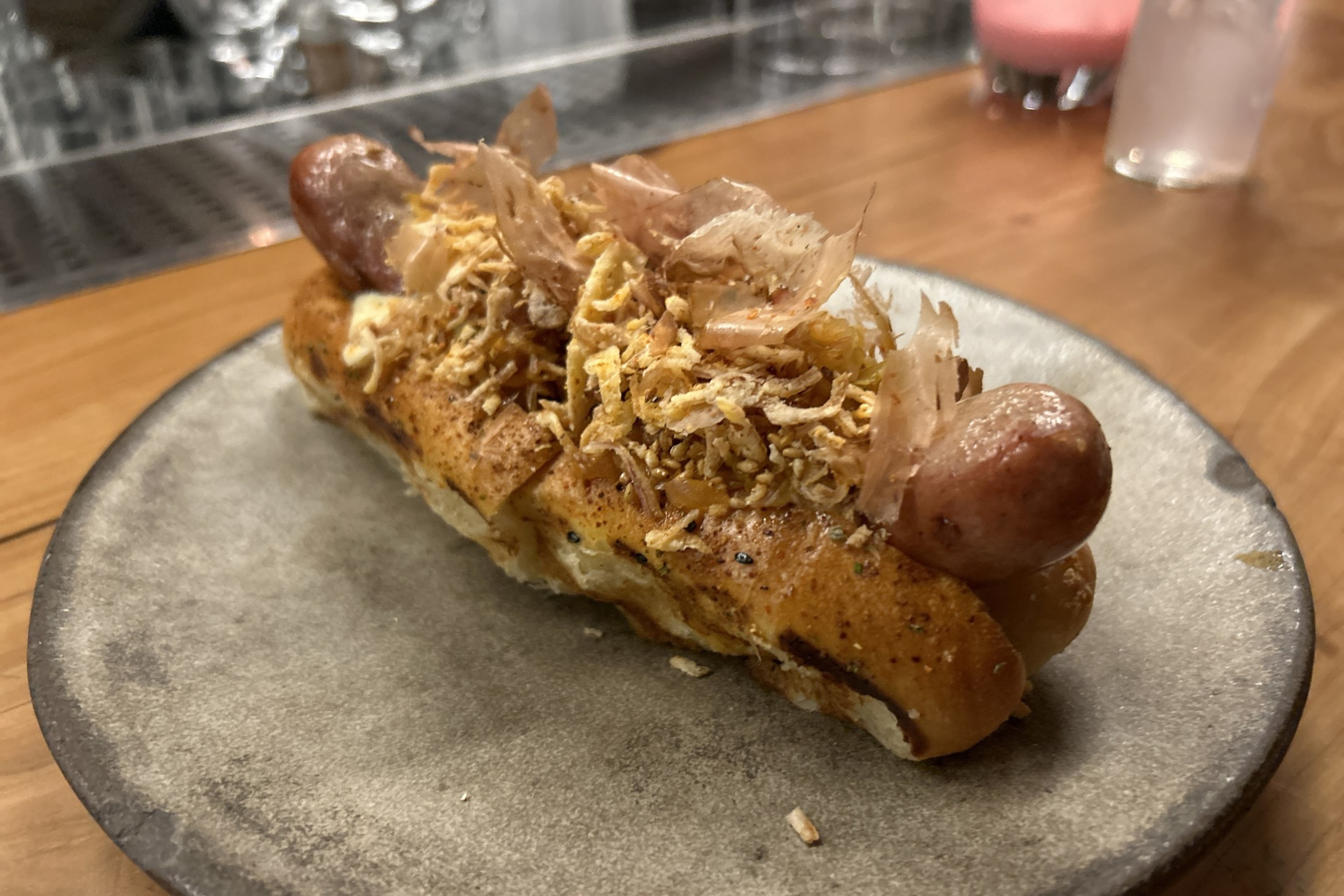 An overhead view of a hotdog topped with kimchi, bonito flakes, toasted sesame, fried shallots and rosemary aioli on a togarashi bun.
