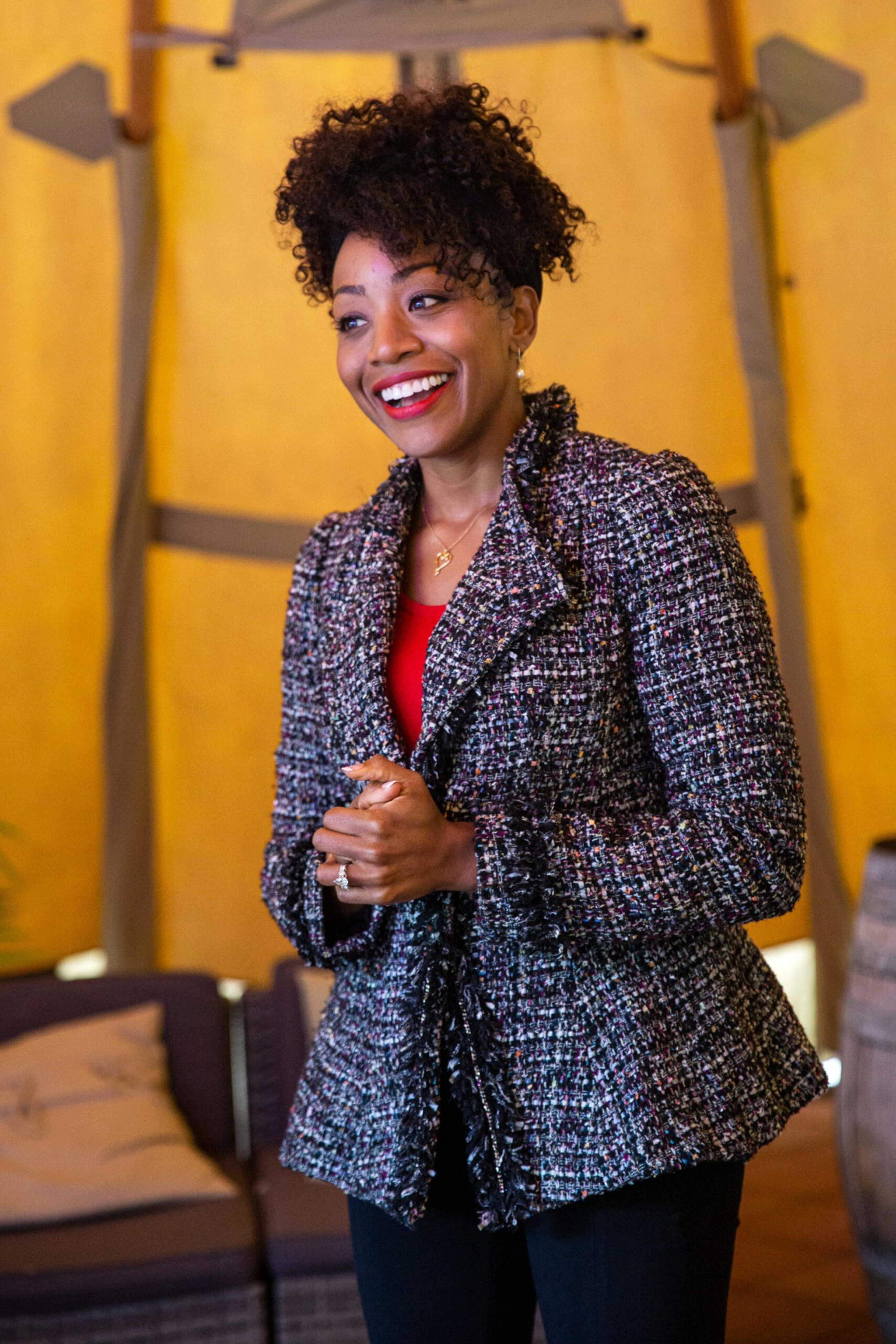 A smiling woman with curly hair wears a textured jacket over a red top, standing indoors with cozy furniture and warm lighting in the background.