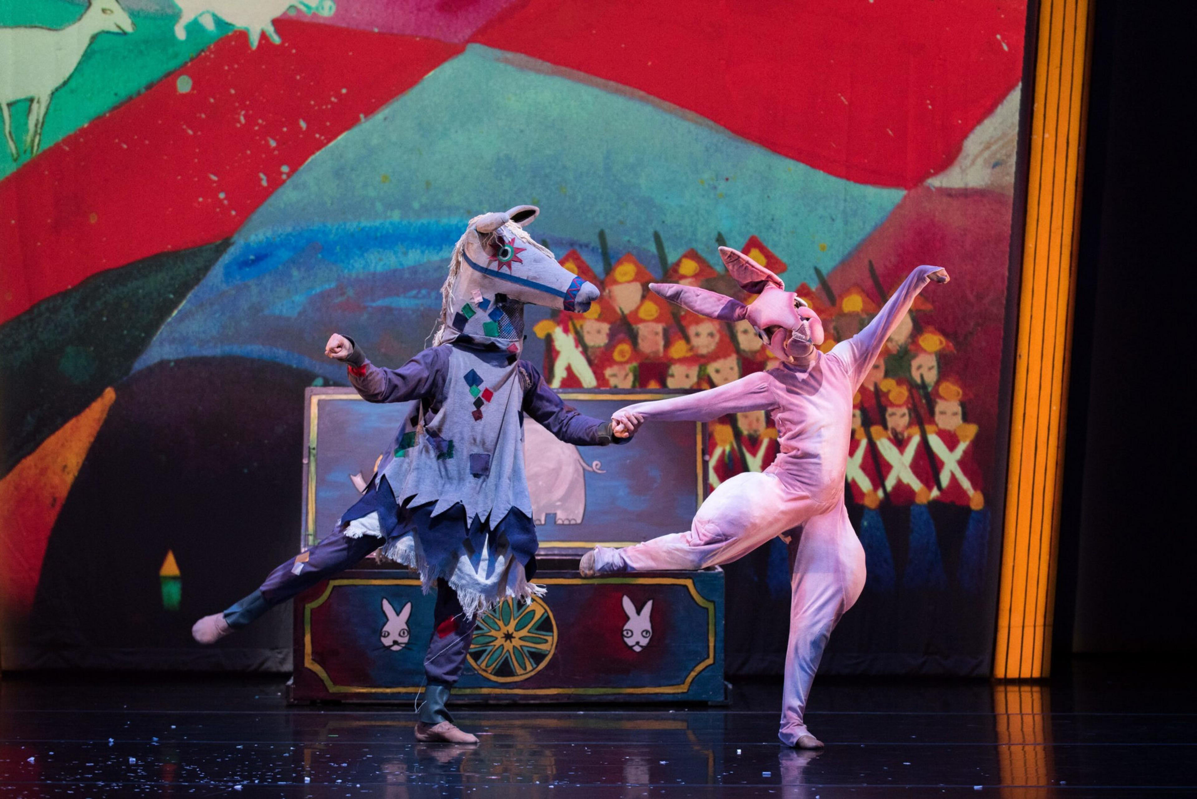 Two performers in animal costumes—one horse and one rabbit—dance on stage with colorful backdrops and toy soldiers painted in the background.