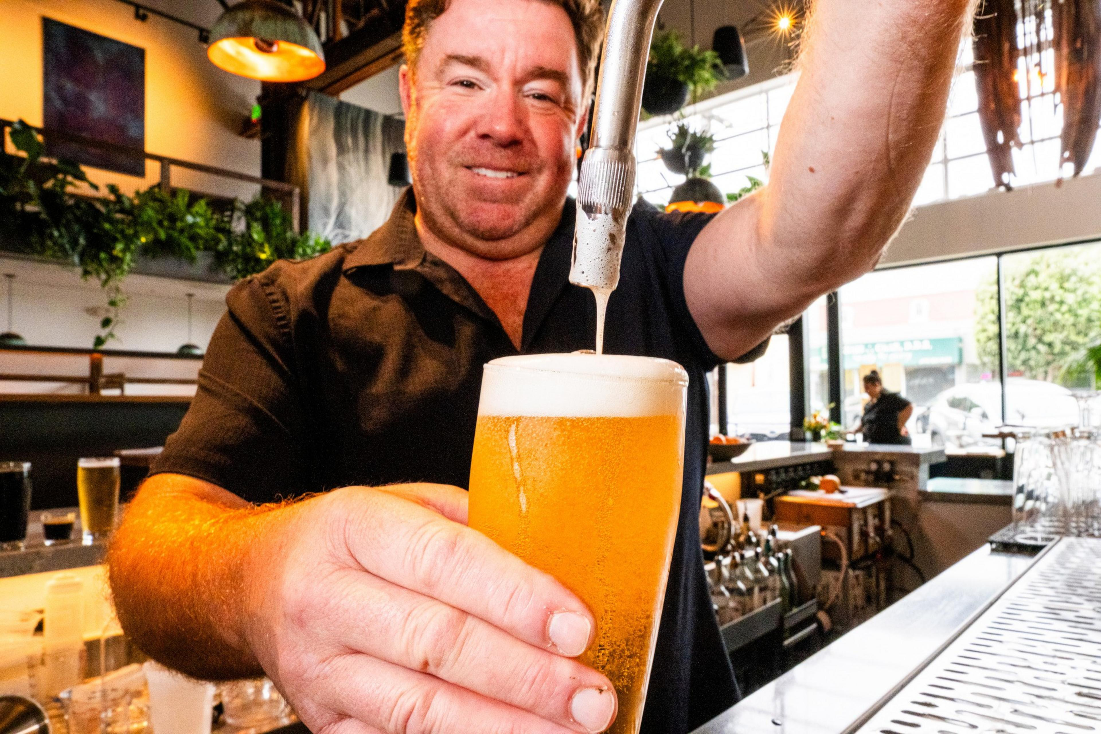 A smiling bartender pours a golden beer with a frothy head from a tap in a bright, modern bar with plants and hanging lights.