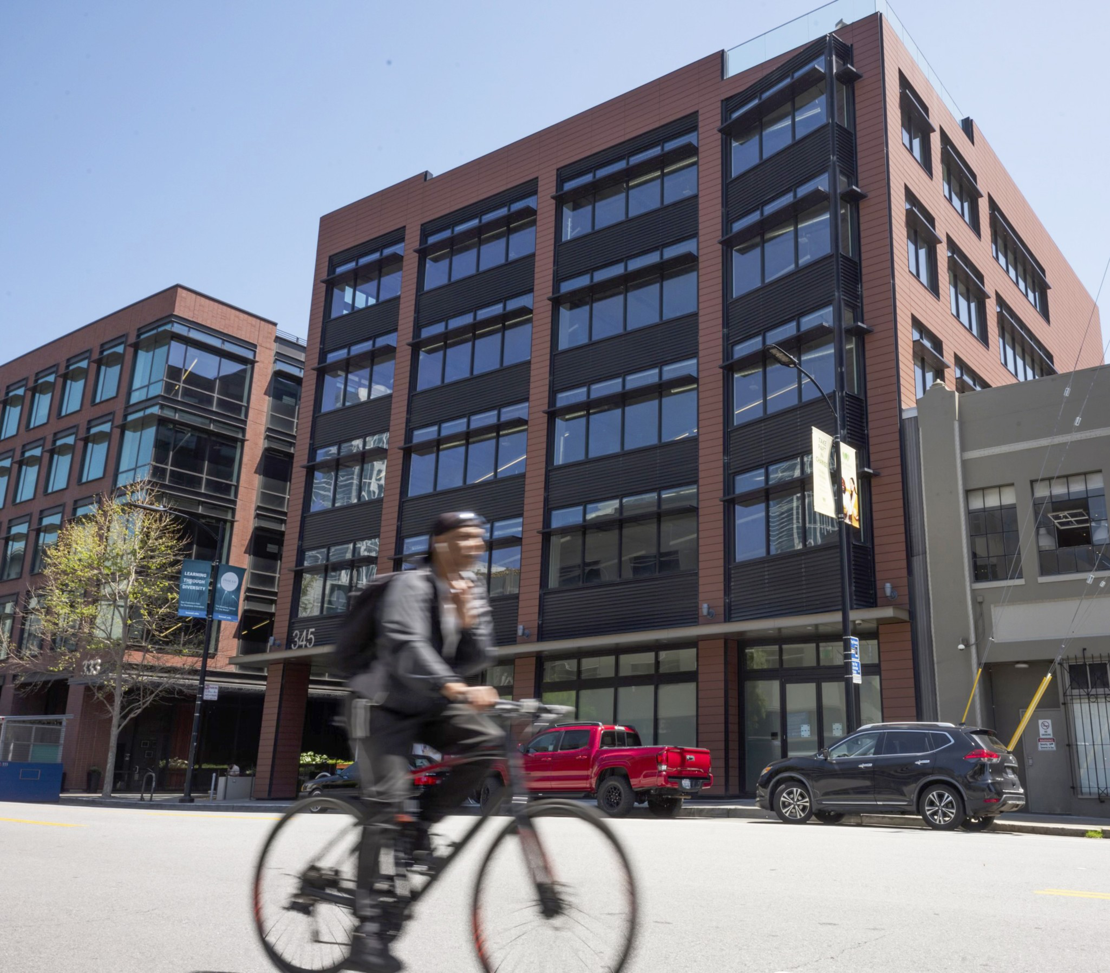 A cyclist in motion blurs past a modern building with large windows and parked cars on a sunny day.