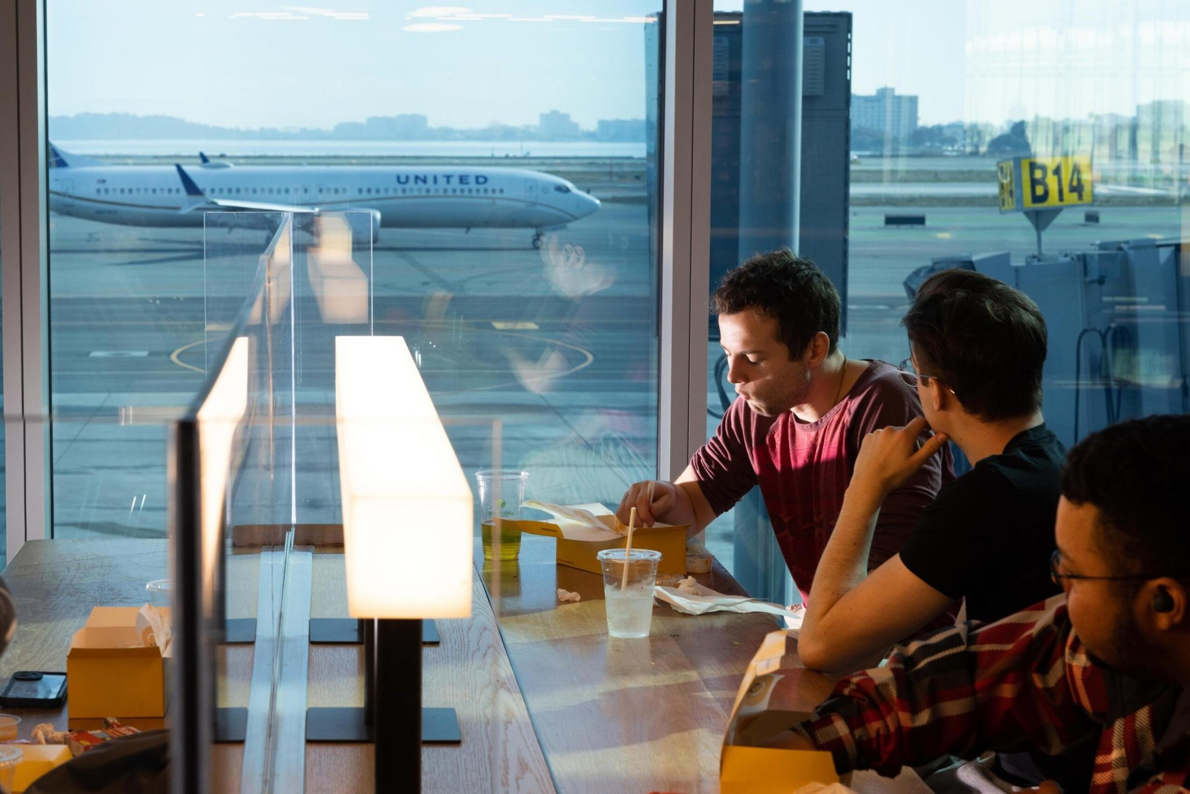 Three people sit at a table by an airport window, eating and drinking, with a United airplane visible outside near gate B14.