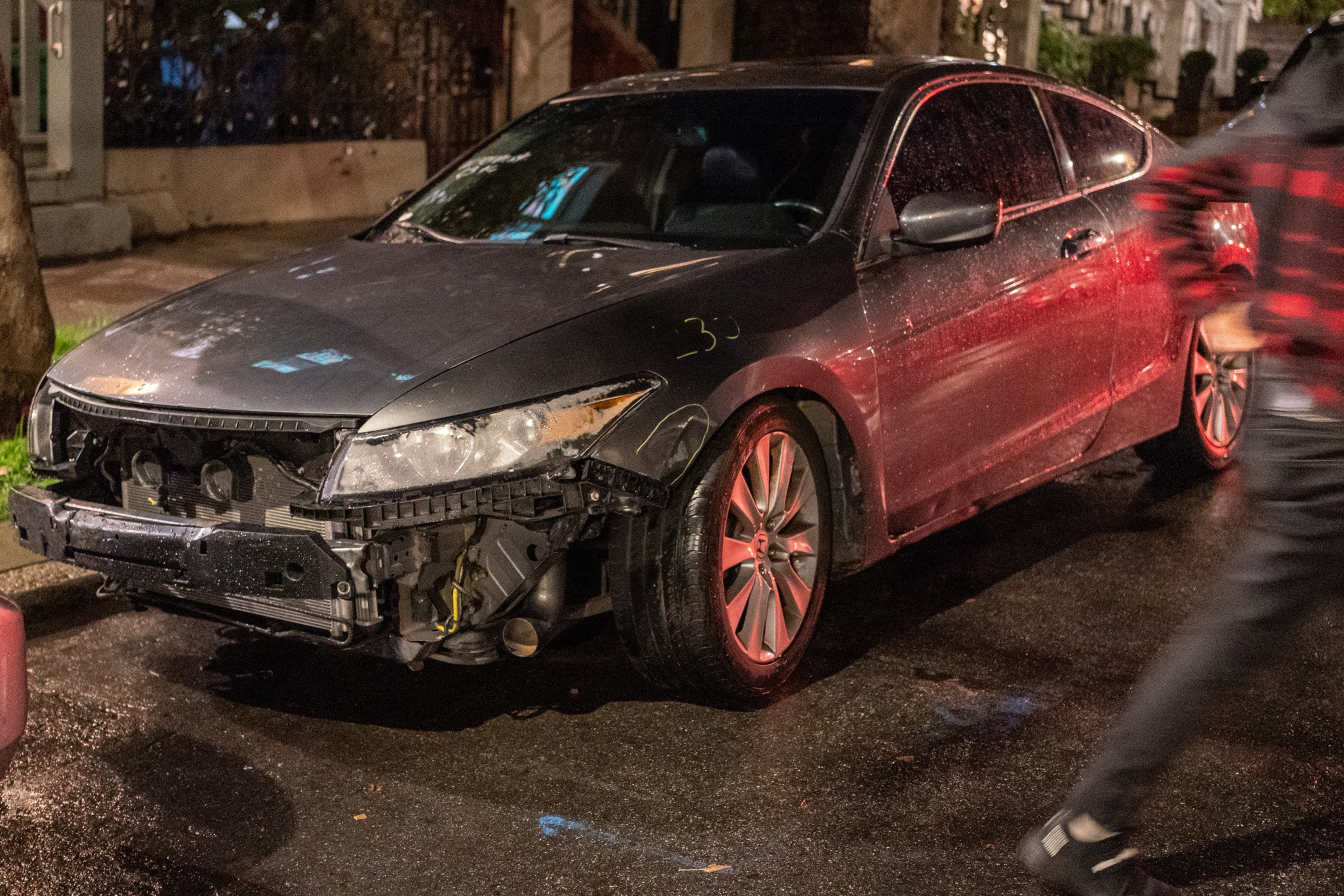 A damaged car with missing front bumper, exposed engine, and a blurred person walking by.