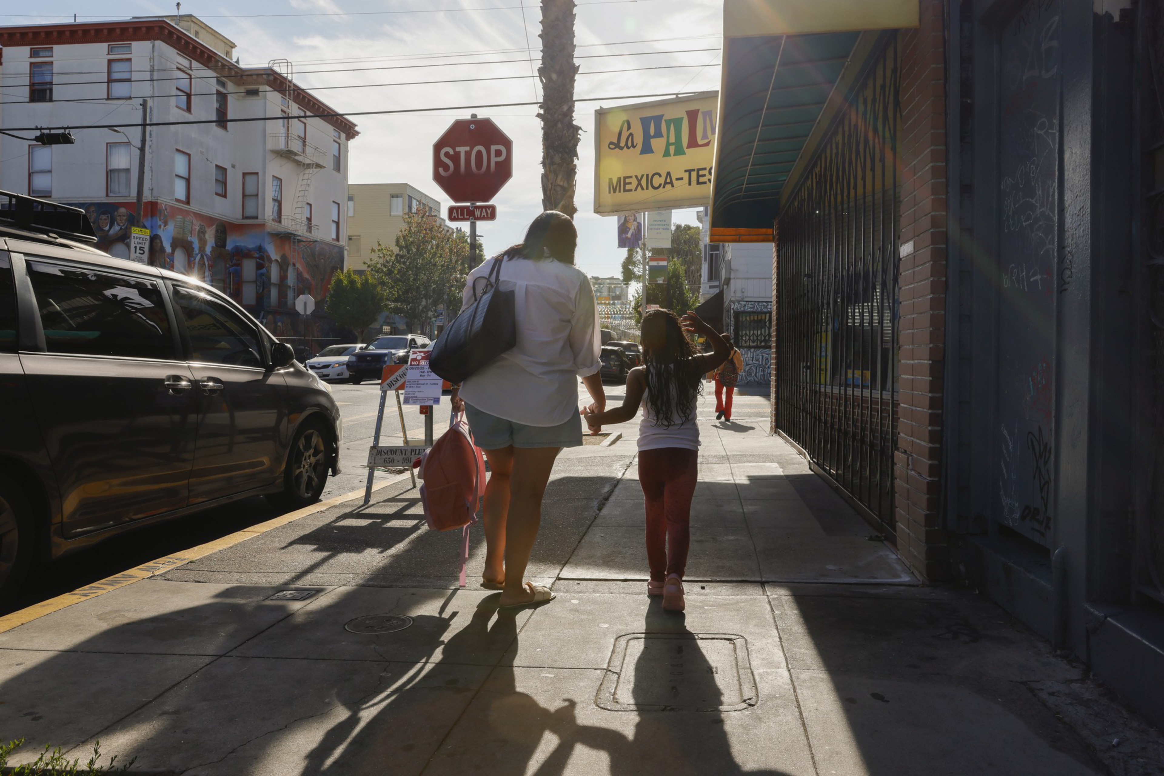 A woman and a child walk hand in hand down a sunny city sidewalk, with a stop sign, parked cars, and a "La Plaza Mexica-Tex" sign visible.