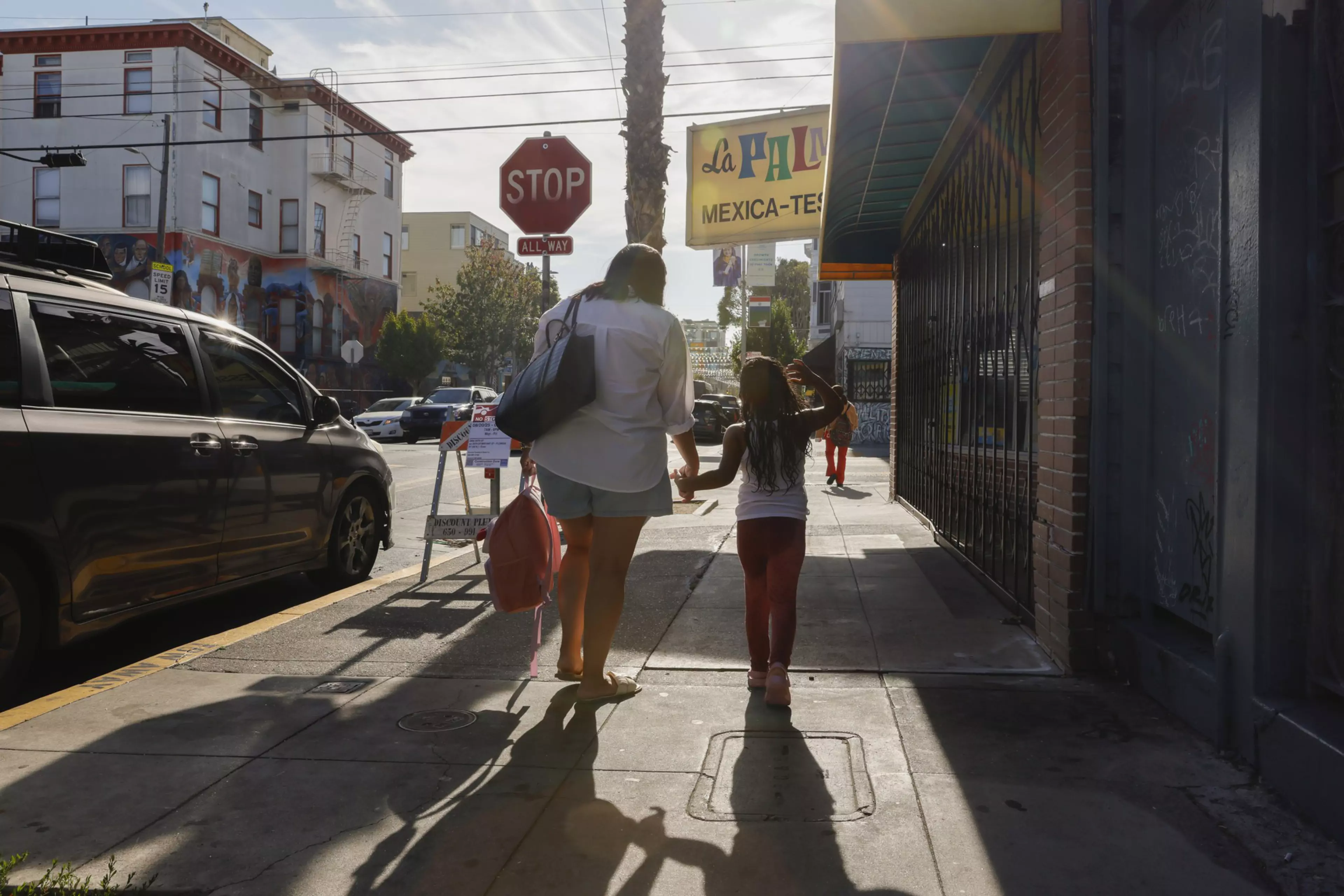 A woman and a child walk hand in hand down a sunny city sidewalk, with a stop sign, parked cars, and a "La Plaza Mexica-Tex" sign visible.