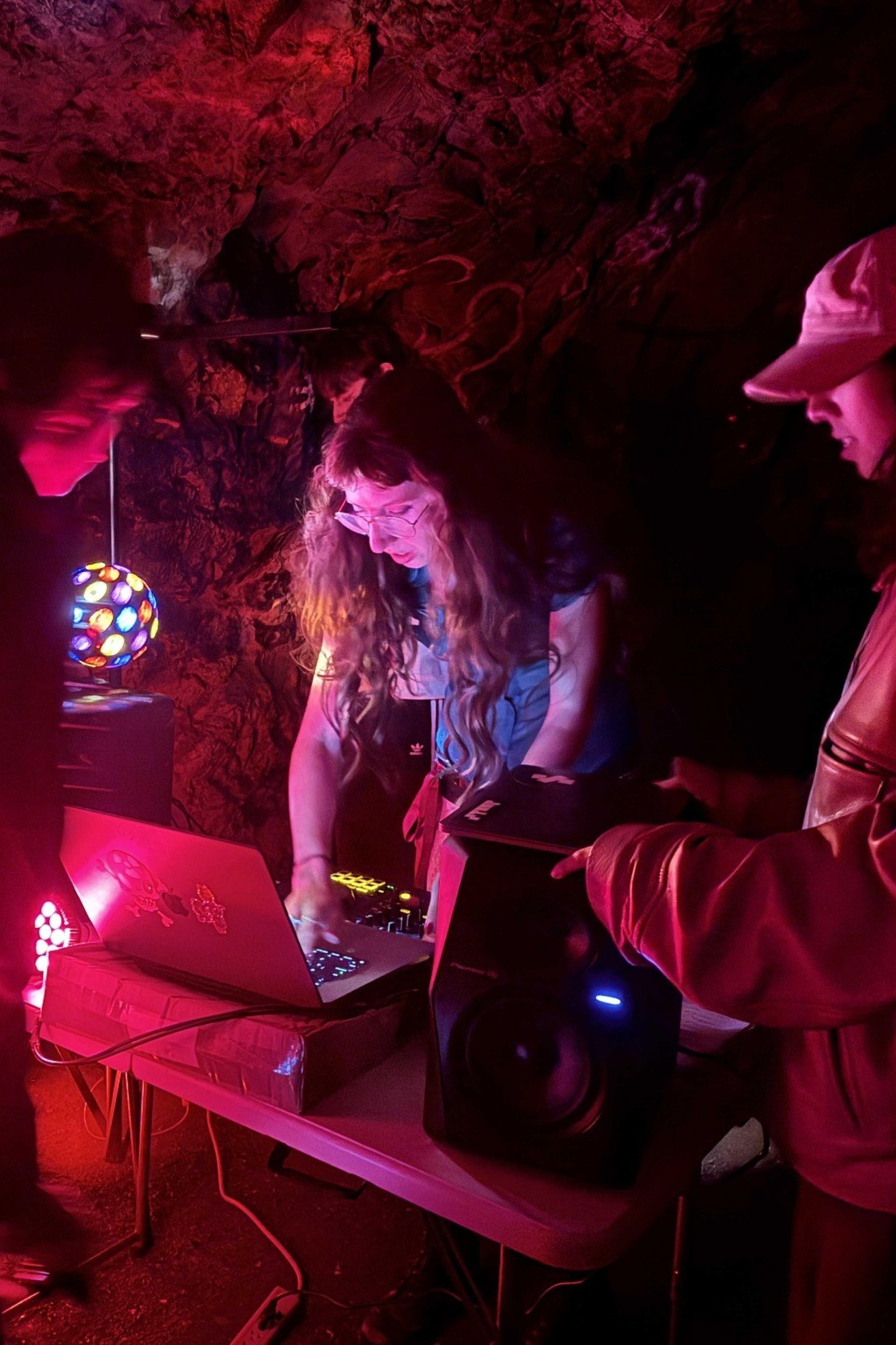 A dimly lit cave features four people gathered around electronic equipment on a table, illuminated by colorful lights, including a disco ball and a glowing red light.