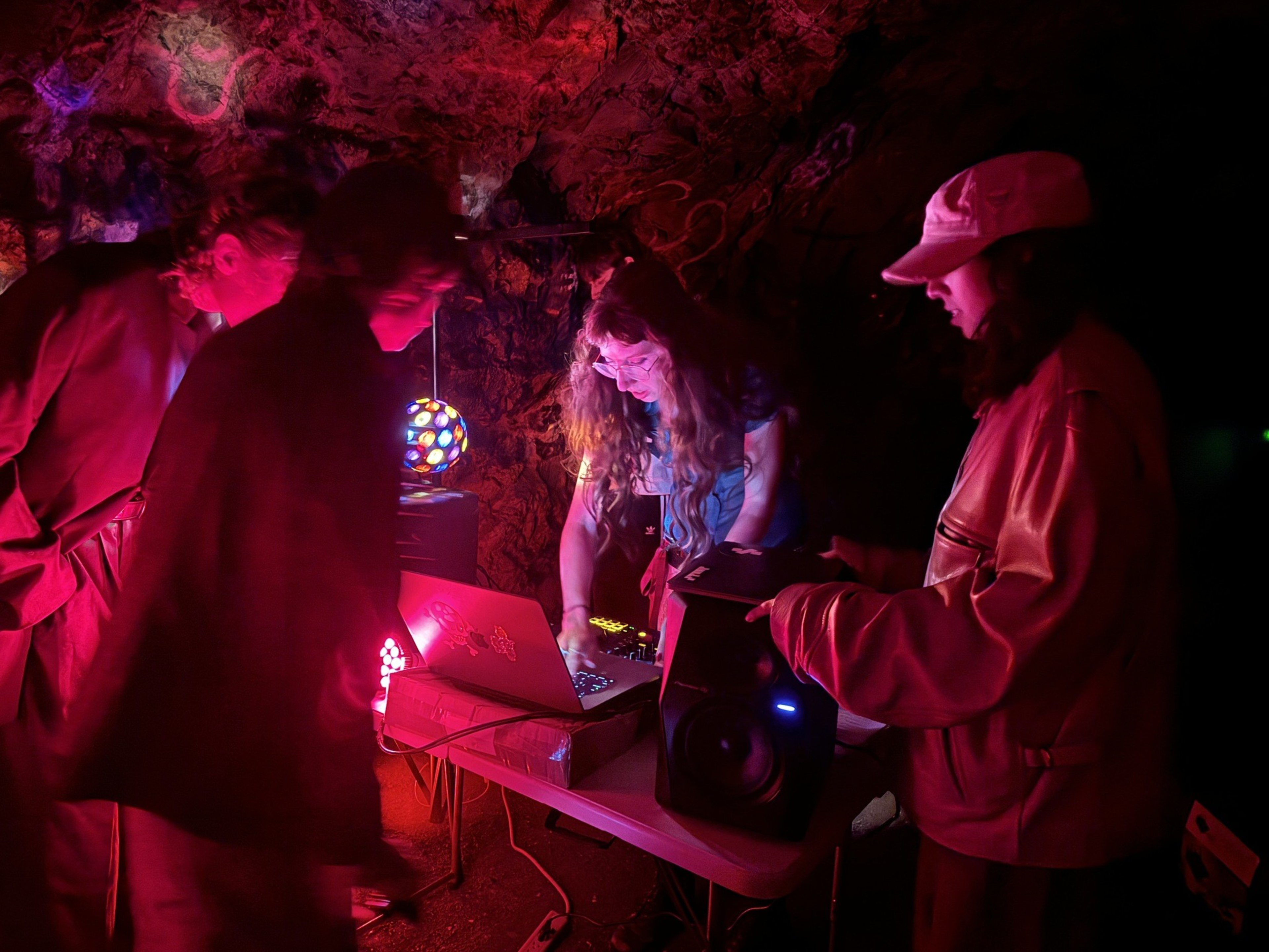 A dimly lit cave features four people gathered around electronic equipment on a table, illuminated by colorful lights, including a disco ball and a glowing red light.