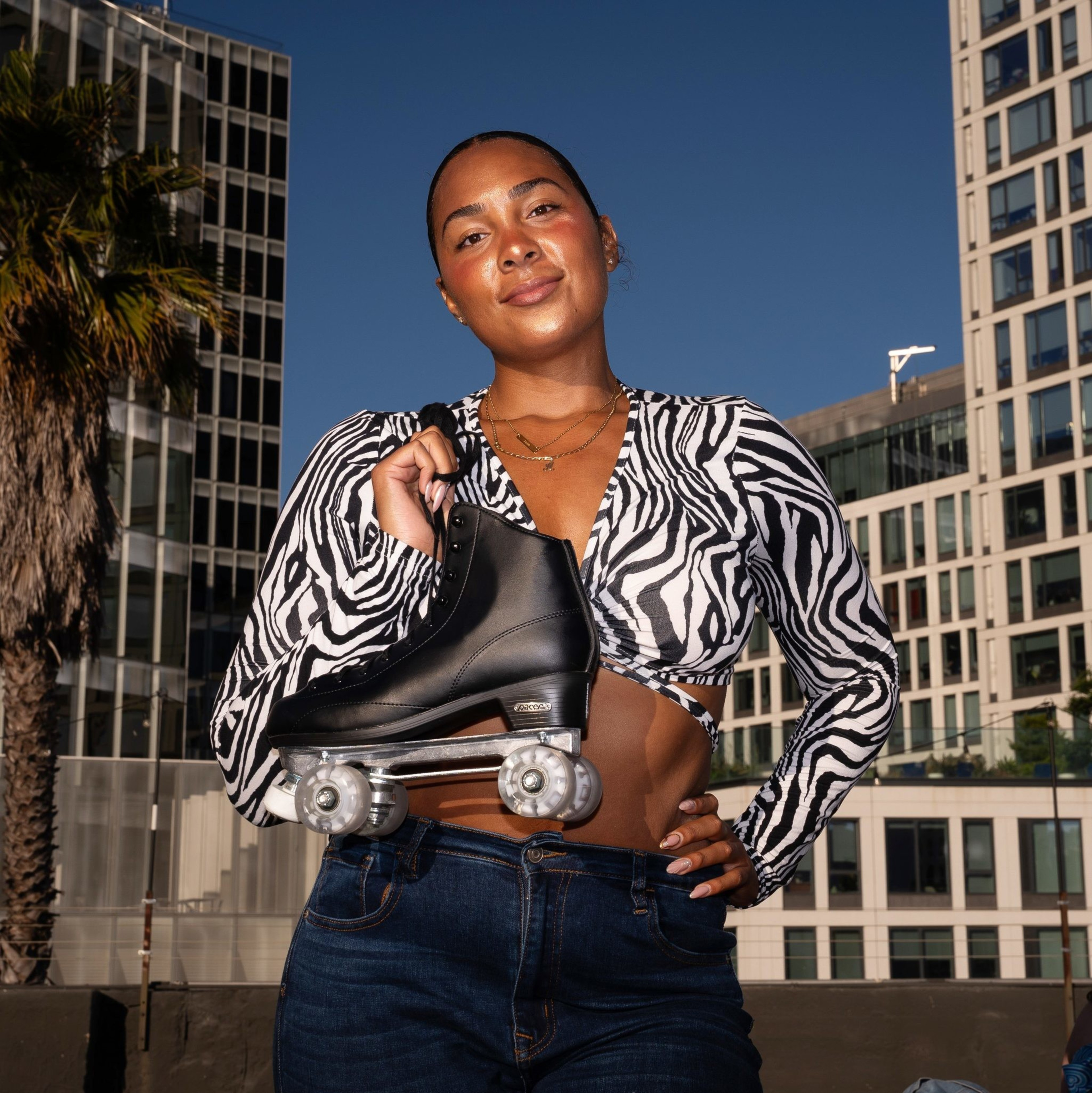 A woman in a zebra-print top and jeans holds a roller skate over her shoulder, standing confidently with skyscrapers and palm trees in the background.