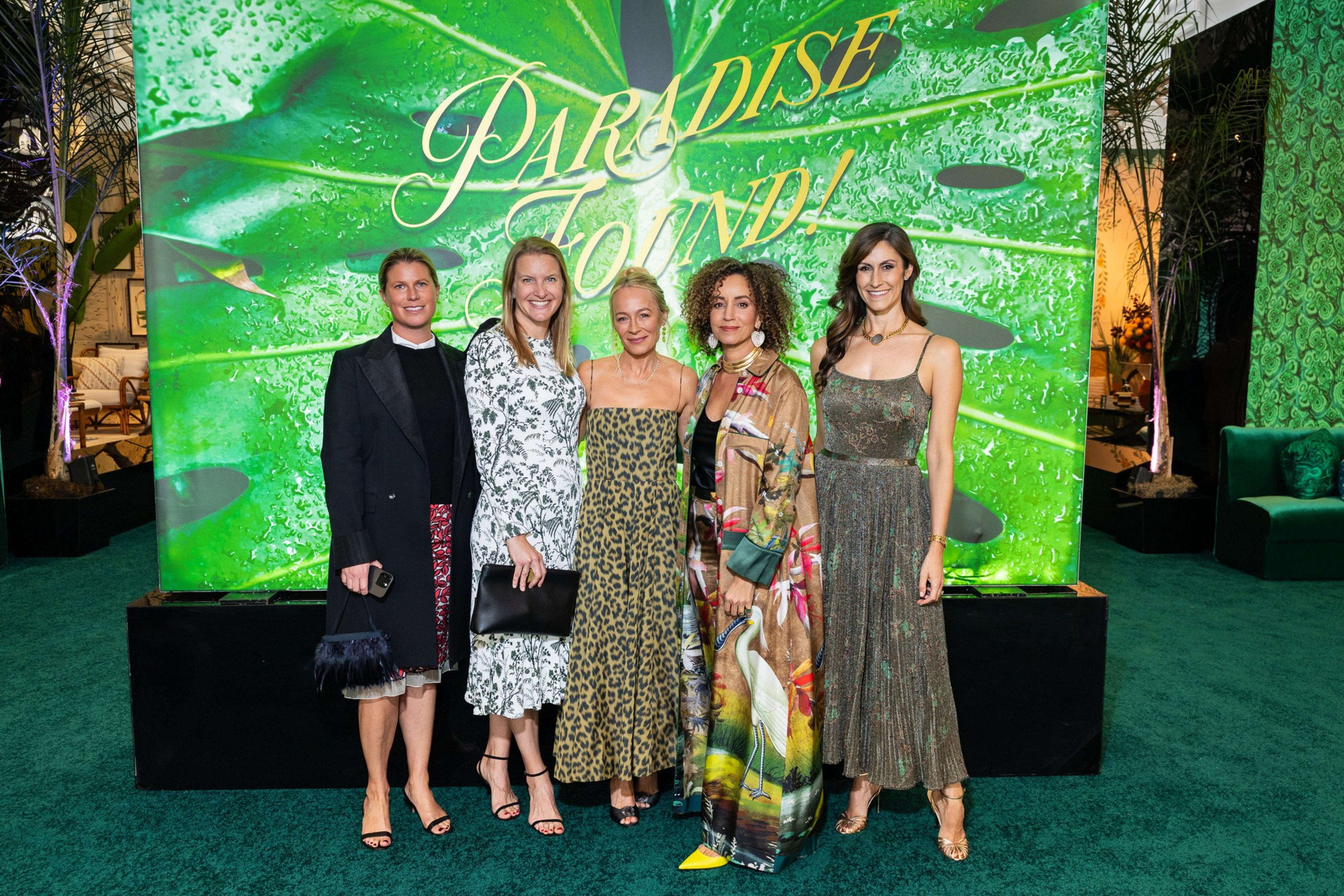 Five women stand on green carpet in front of a large green leafy backdrop with the words “PARADISE FOUND” in gold lettering.