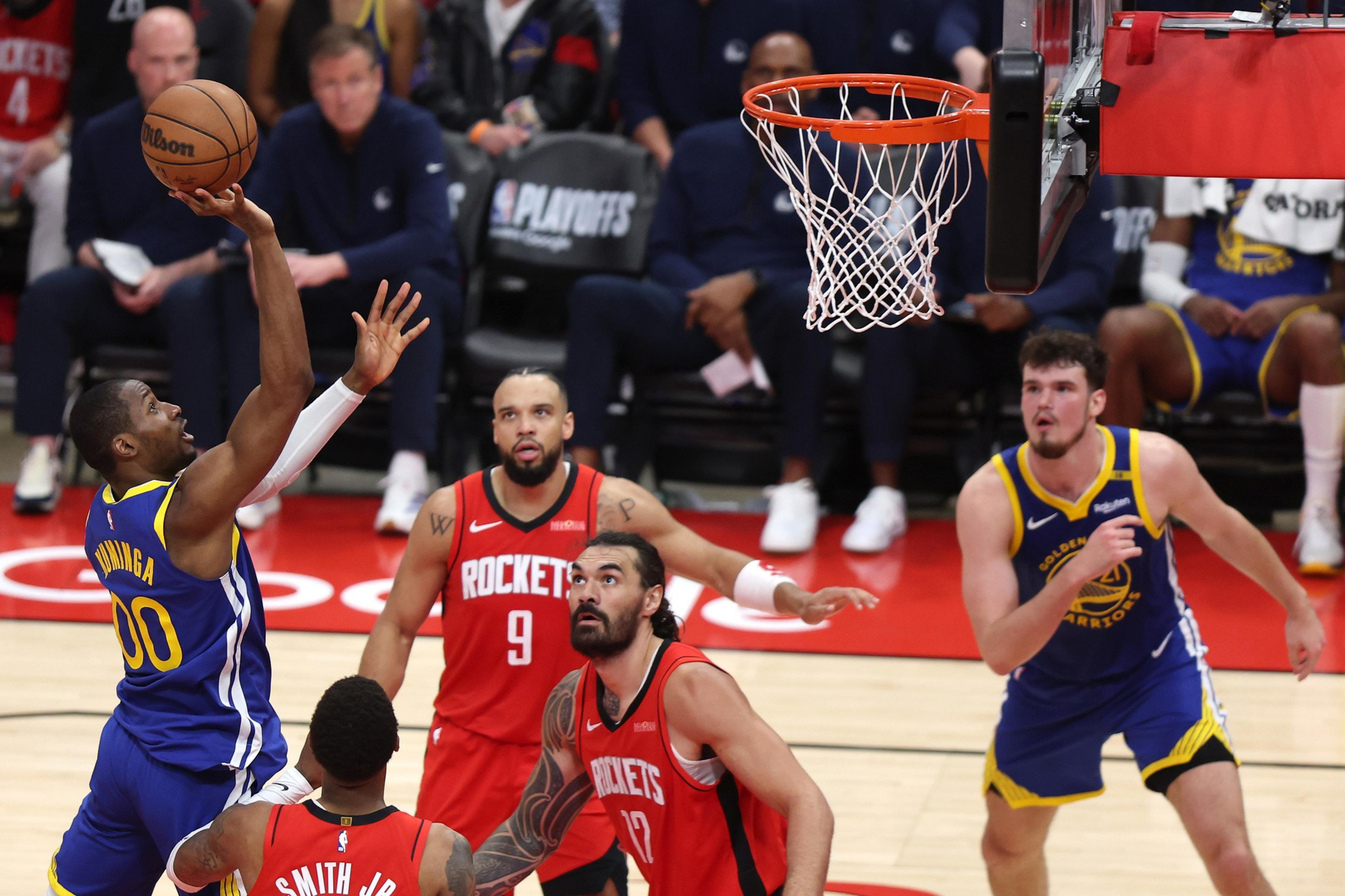 A basketball player in a blue jersey is attempting a shot. Opposing players in red jerseys watch, with one jumping to defend. The scene is intense and action-packed.