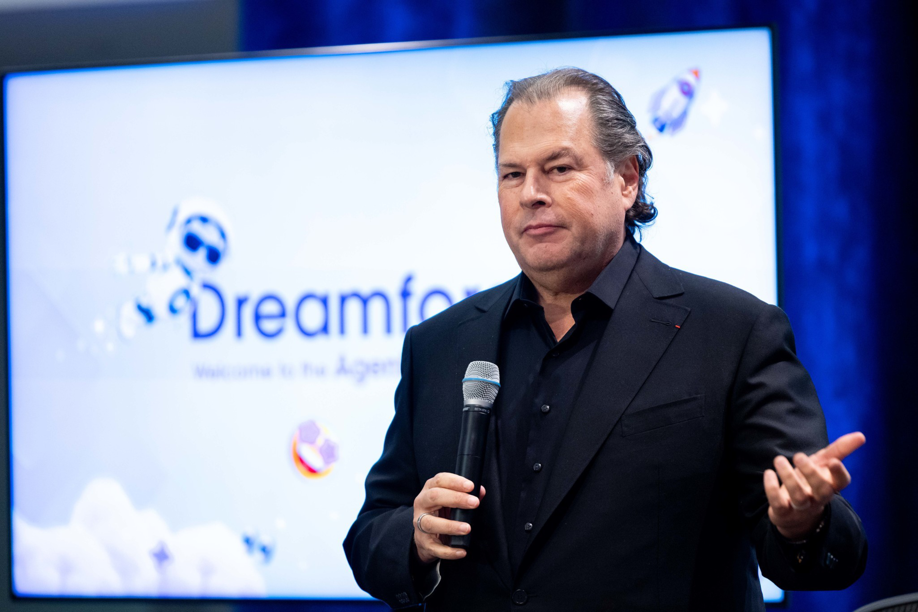A man in a black suit holds a microphone and gestures with his hand while speaking in front of a screen displaying the word "Dreamforce."