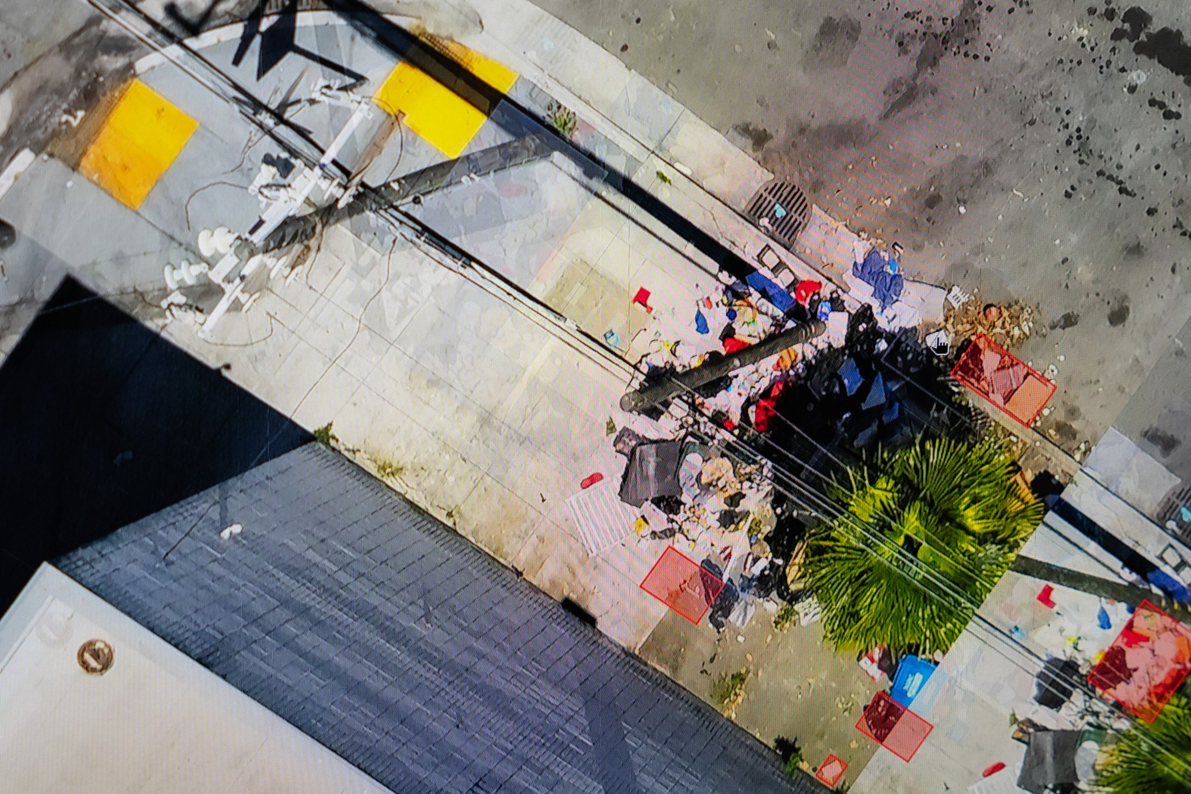 A sidewalk and street corner cluttered with scattered trash, debris, a palm tree, electrical wires, and shadows from nearby buildings.