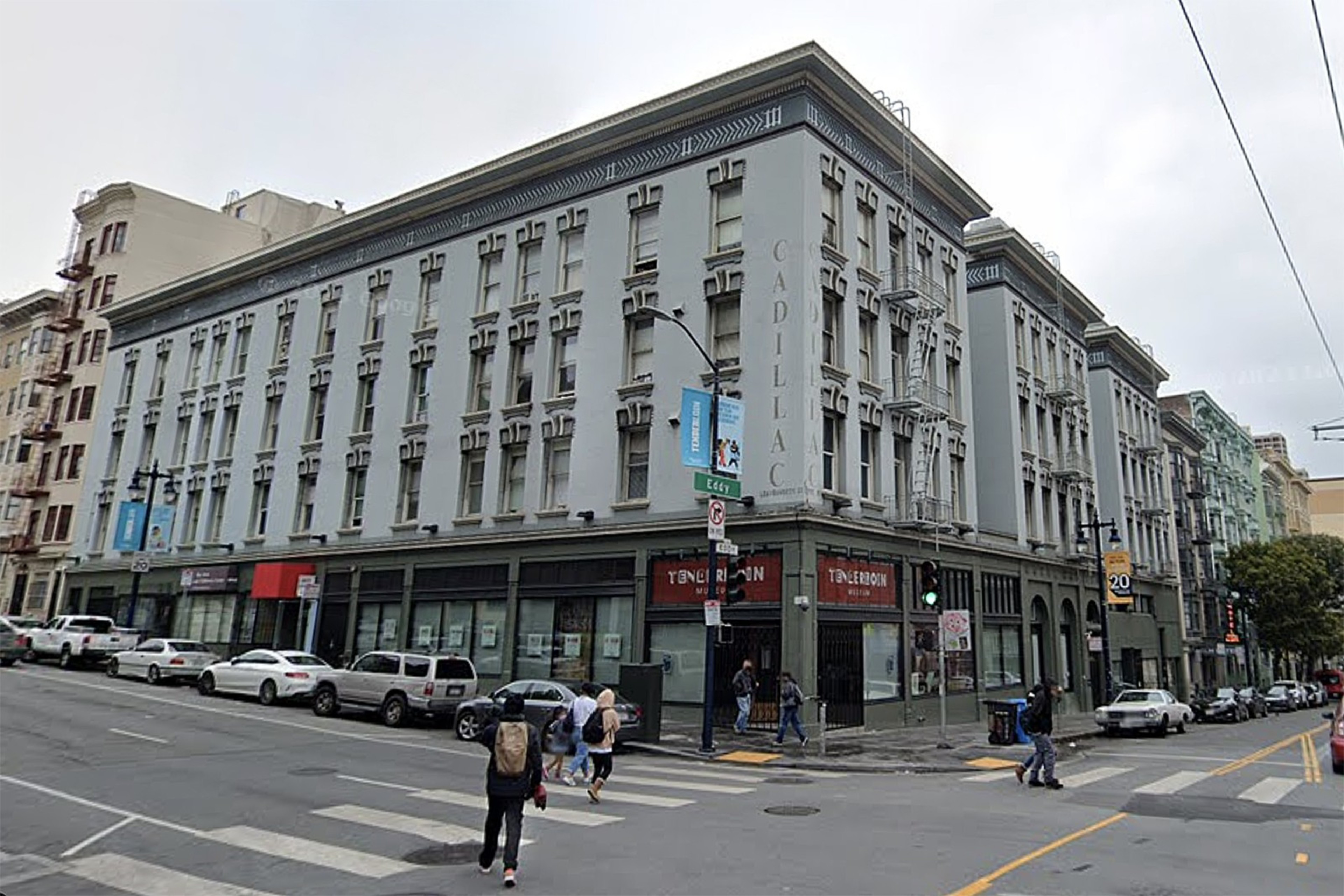 A corner building with rows of windows and ground-floor shops, pedestrians on the sidewalk, and cars parked along the street.