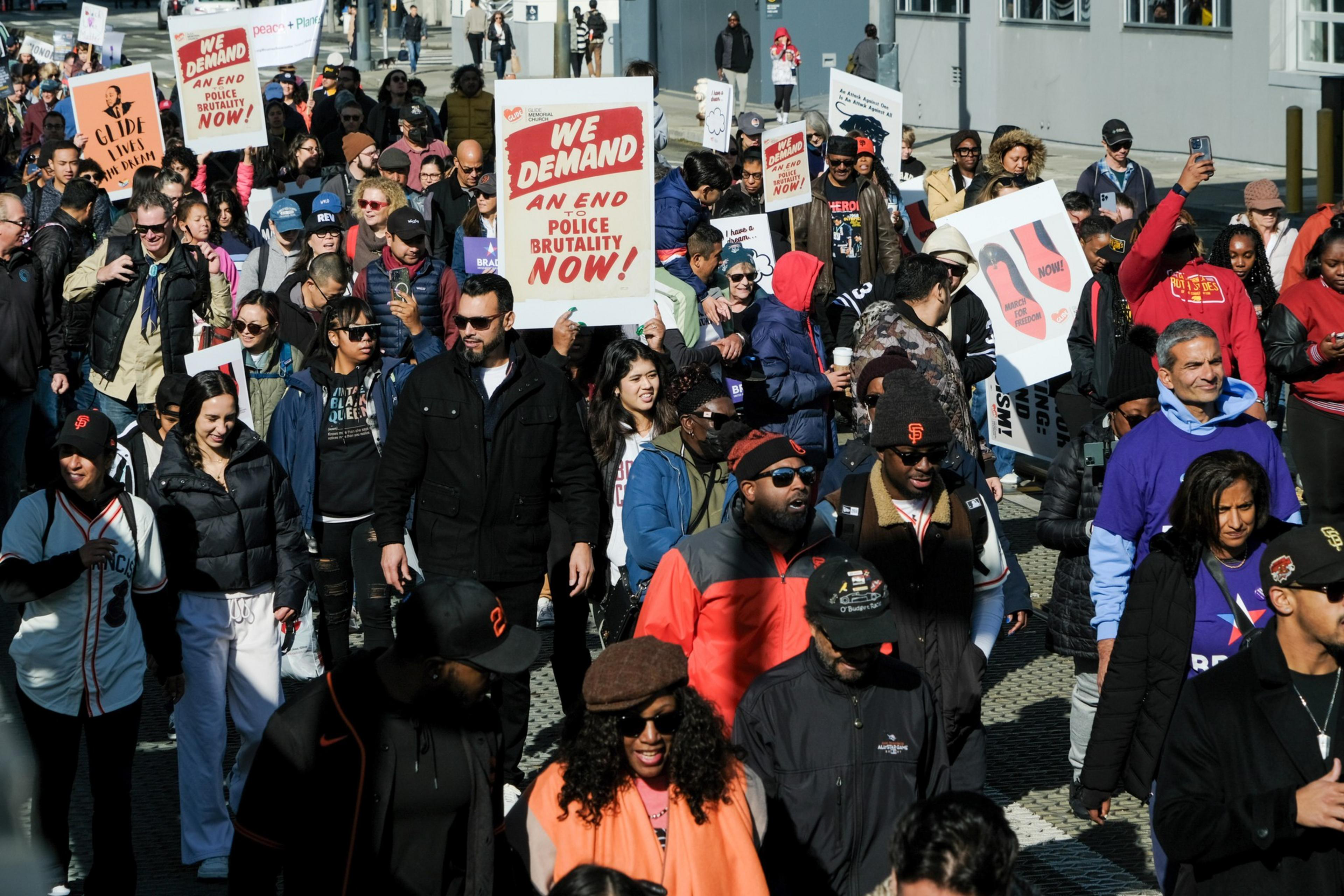 A diverse crowd of protesters holding signs calling for an end to police brutality, moving through a sunny street.