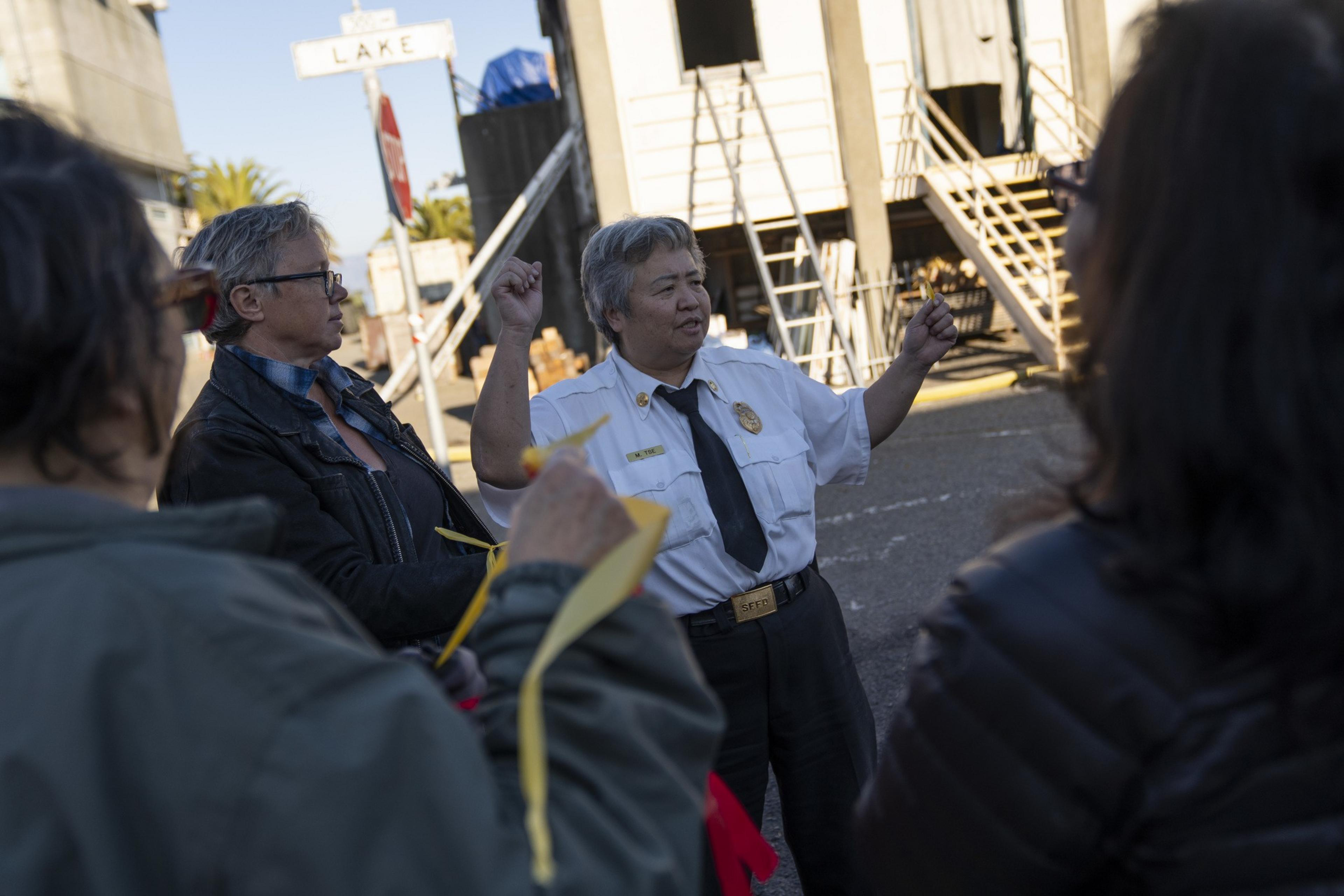 A person in a fire department uniform speaks to a group on a city street. They gesture with both hands, standing in front of a building with staircases.