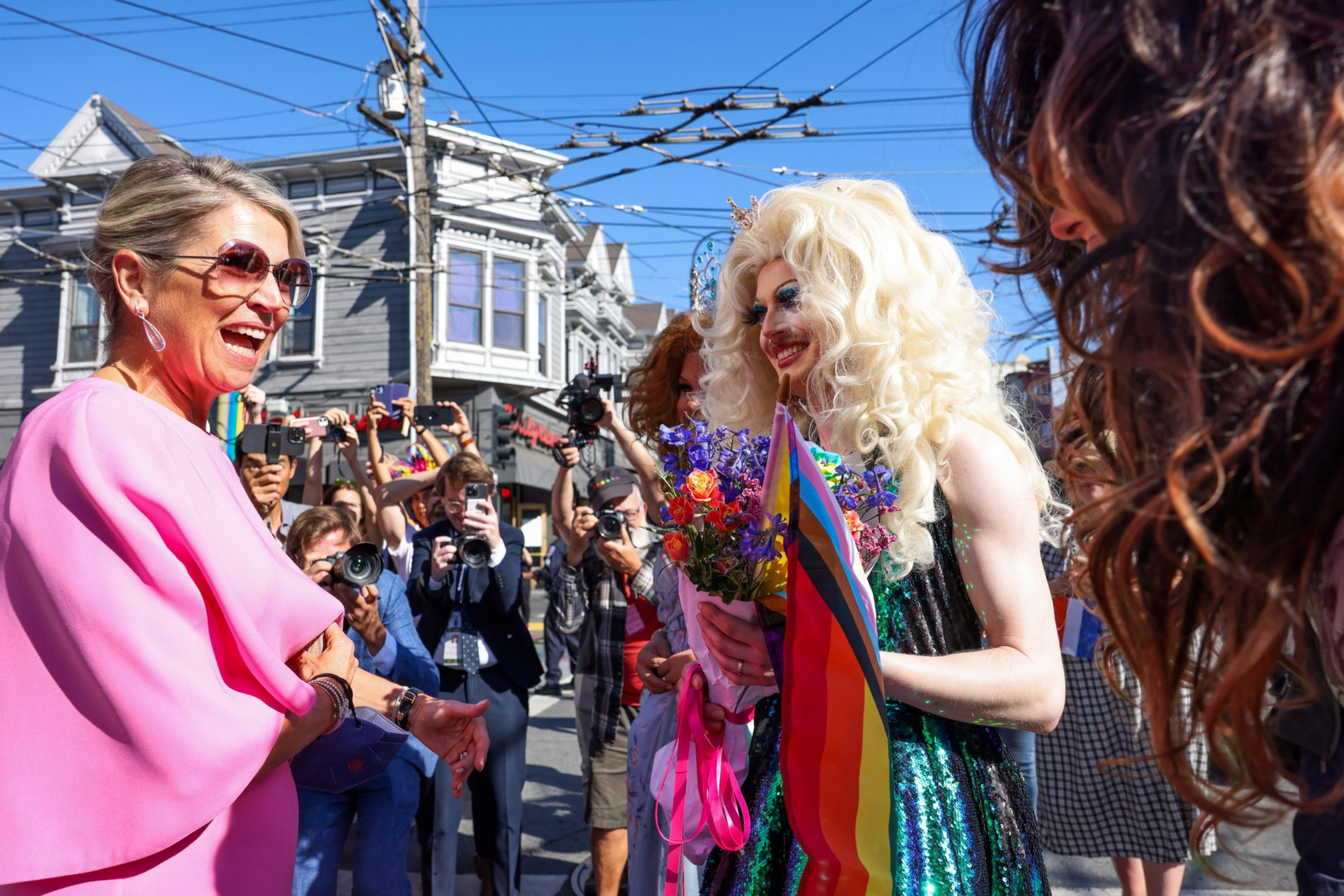 A person in a pink outfit laughs while shaking hands with a drag queen holding flowers and a rainbow flag, surrounded by photographers and onlookers.