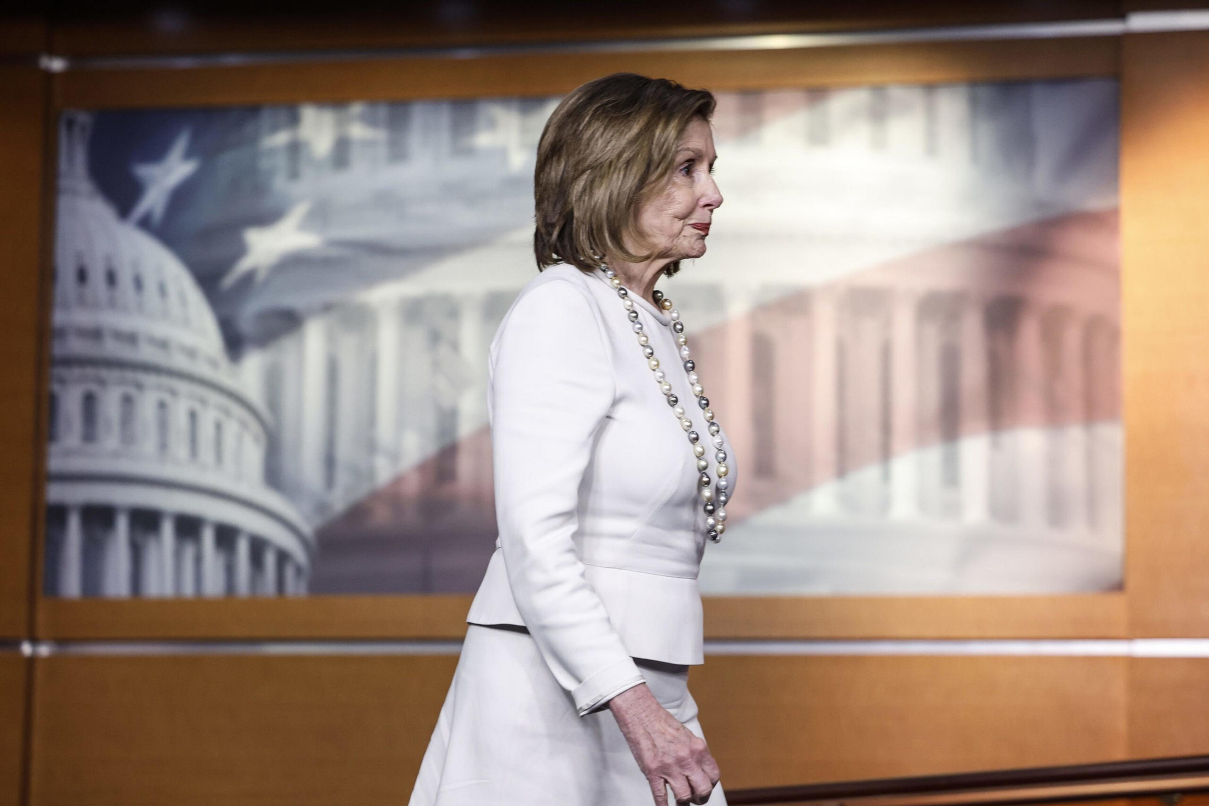 A woman in a white suit and long pearl necklace walks past a backdrop featuring a faded American flag and the U.S. Capitol building.