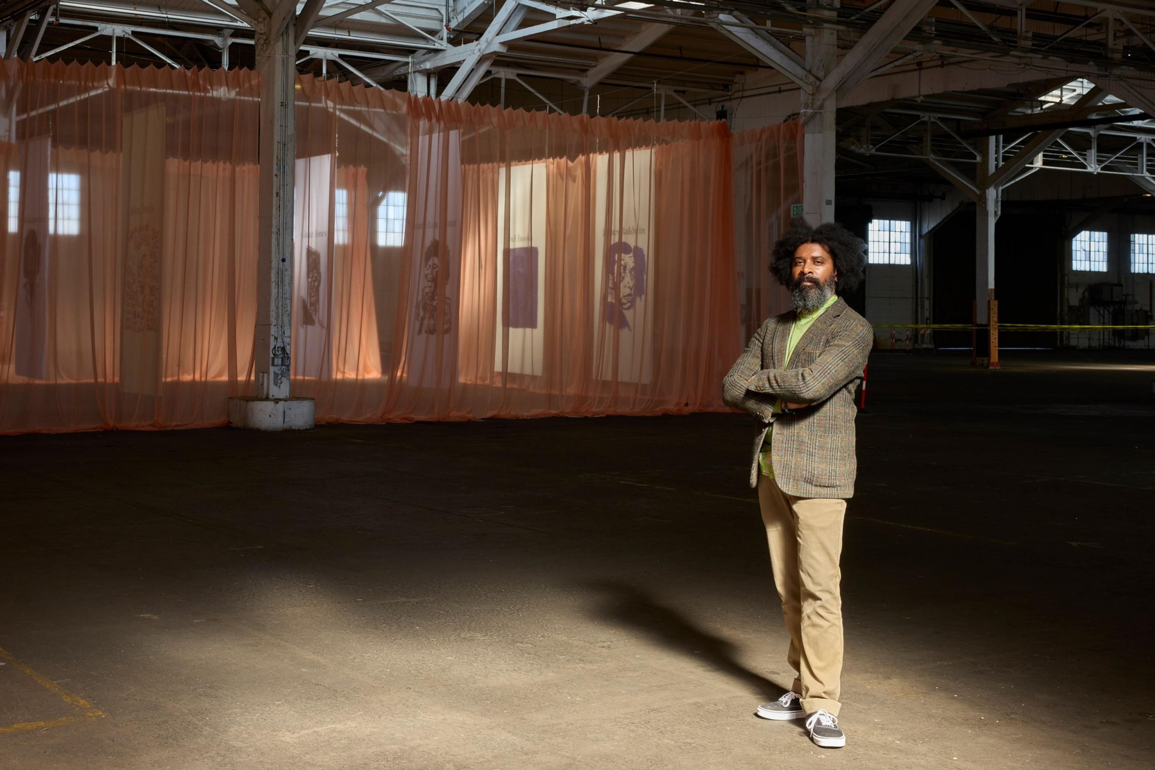 A man with curly hair and a beard stands confidently with arms crossed in a large, dimly lit warehouse with light pink translucent curtains displaying faces.