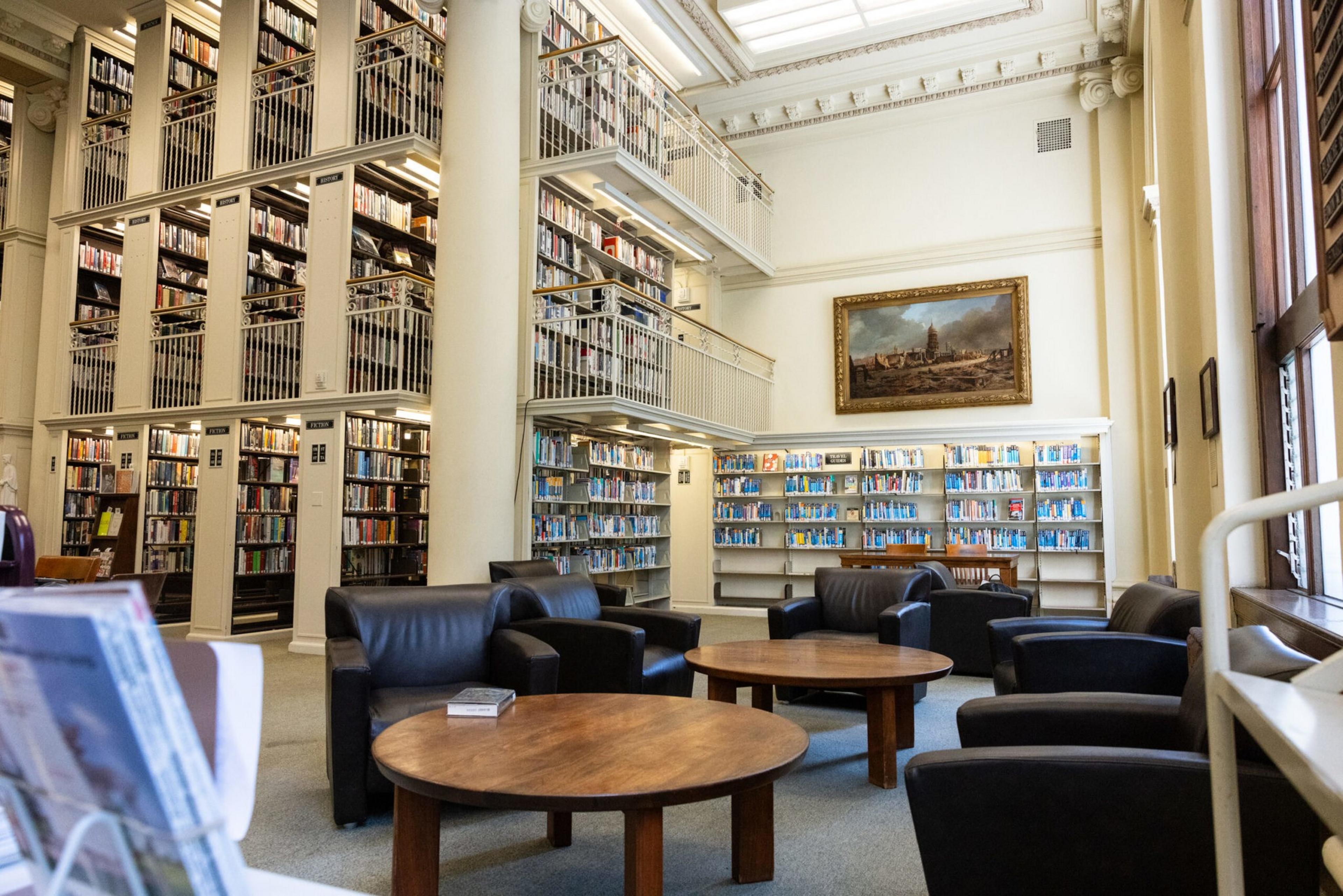A spacious library with tall bookshelves, black armchairs, round wooden tables, and a large framed painting on the wall. Large windows let in natural light.