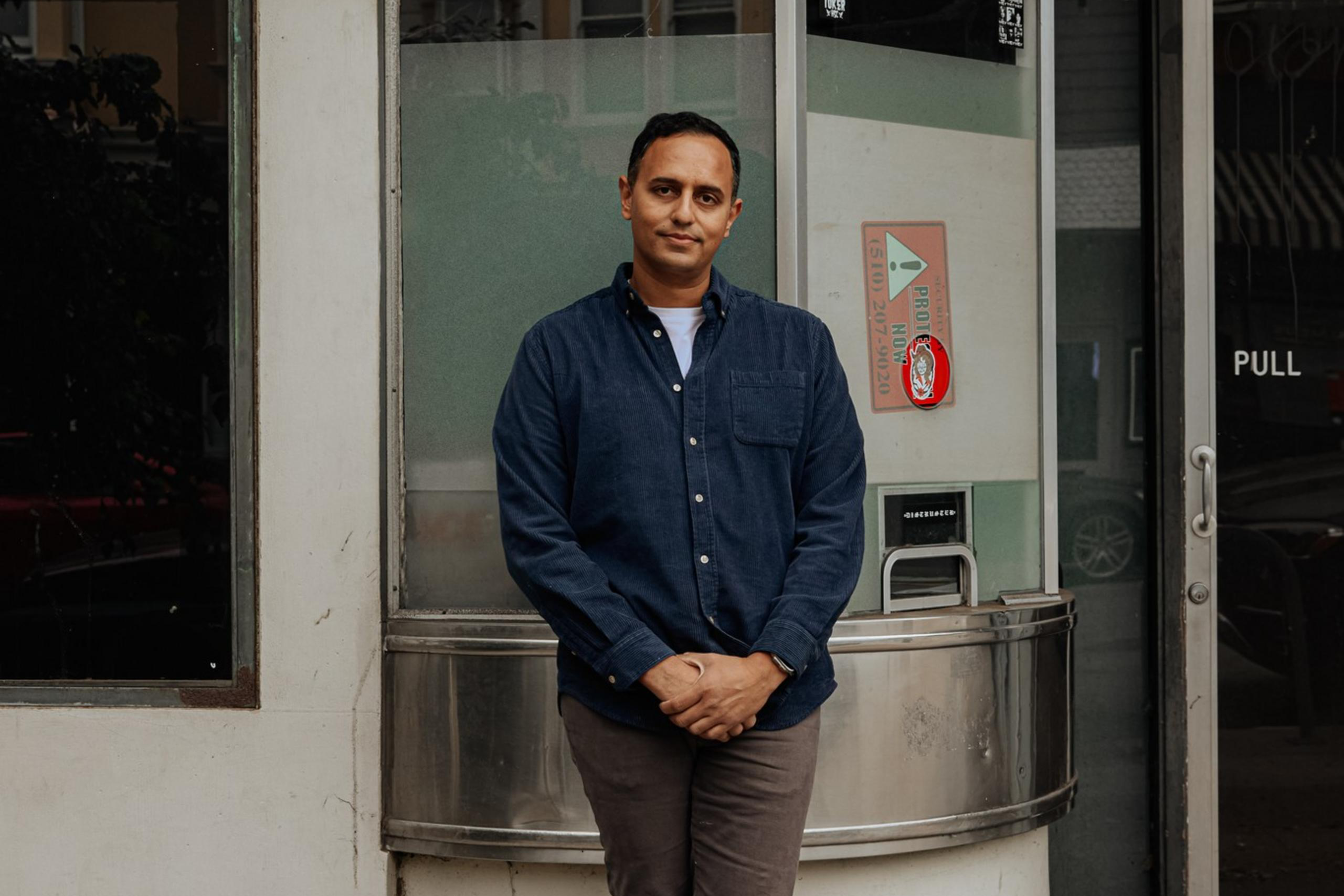 A person in a blue shirt and gray pants stands casually by a rounded corner of a building facade, next to a glass door and a metallic mailbox, with fallen leaves on the ground.