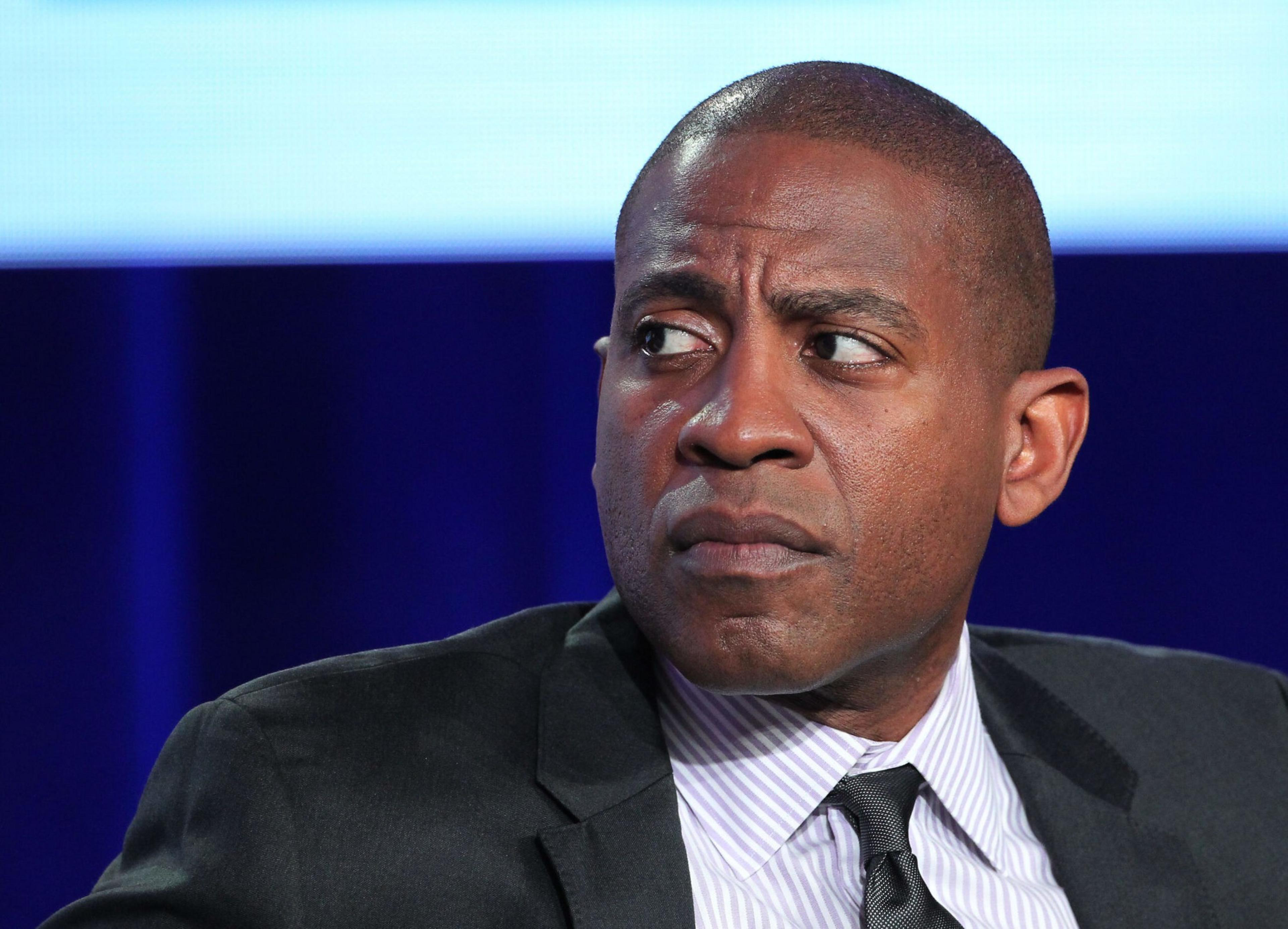 A man in a suit and striped shirt looks to the side with a serious, focused expression against a blurred blue and white background.