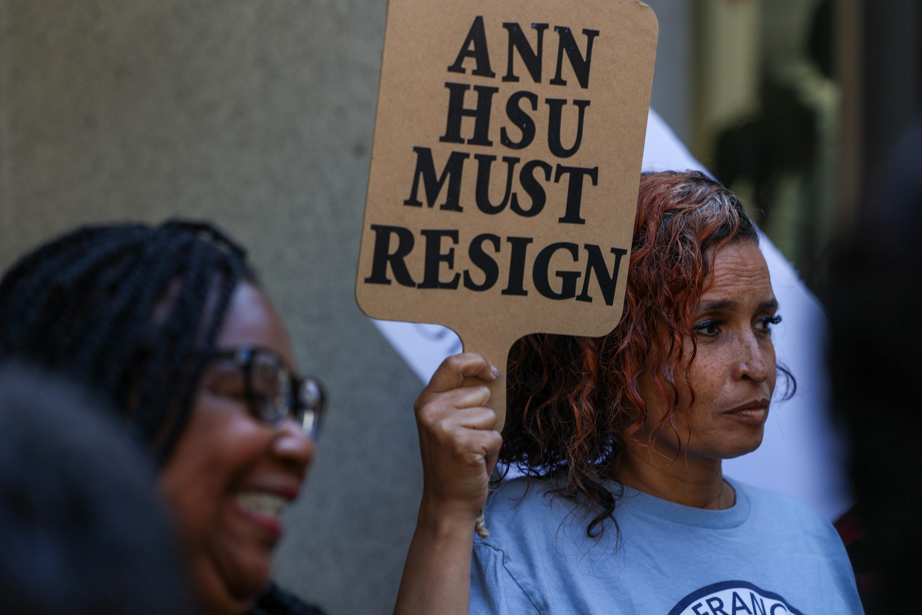 A woman with curly hair holds a sign that reads "ANN HSU MUST RESIGN" while another woman wearing glasses smiles nearby.