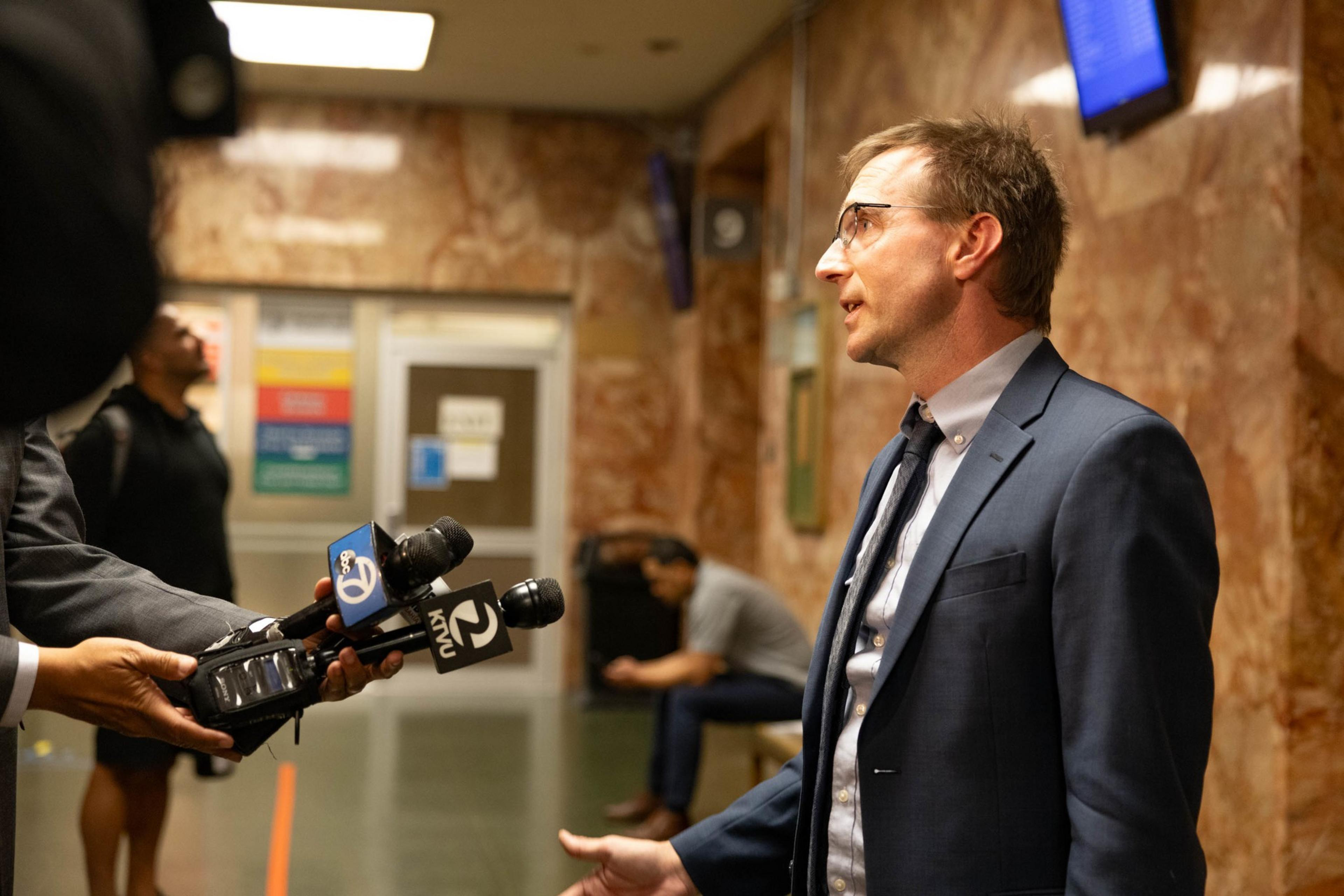 A man in a suit and tie speaks to reporters holding microphones in a marble-walled hallway, while a person sits on a bench in the background.