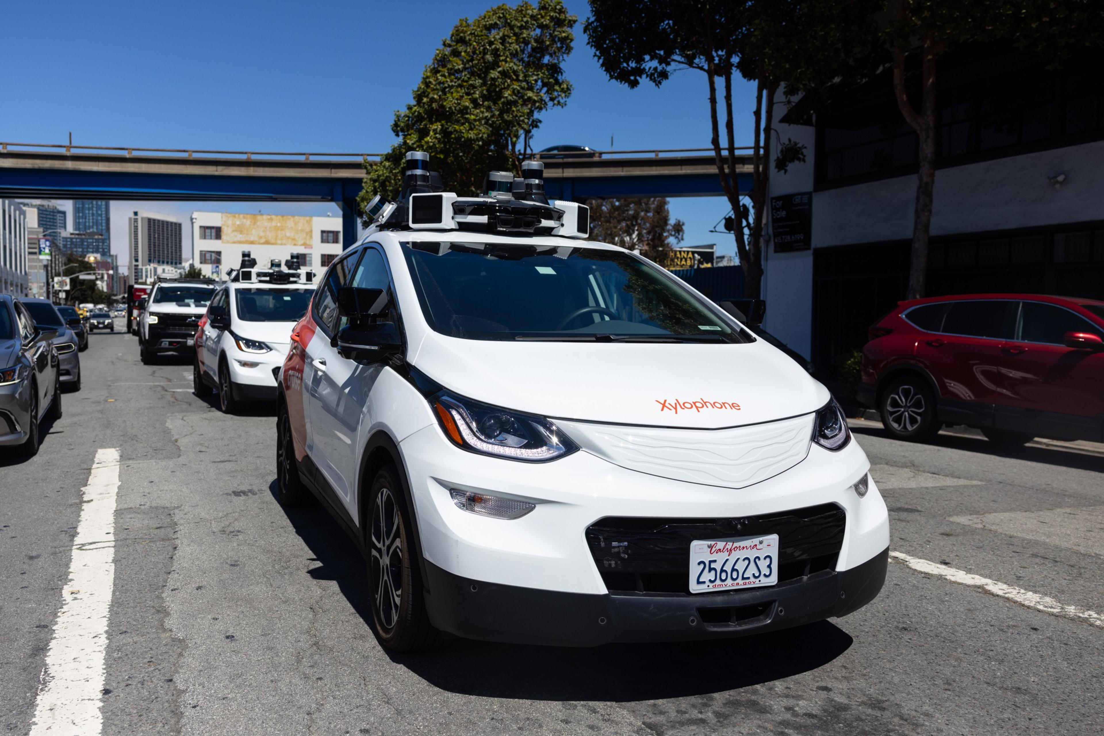 A small white car with a camera display atop its roof waits in traffic at a stoplight on a sunny day.