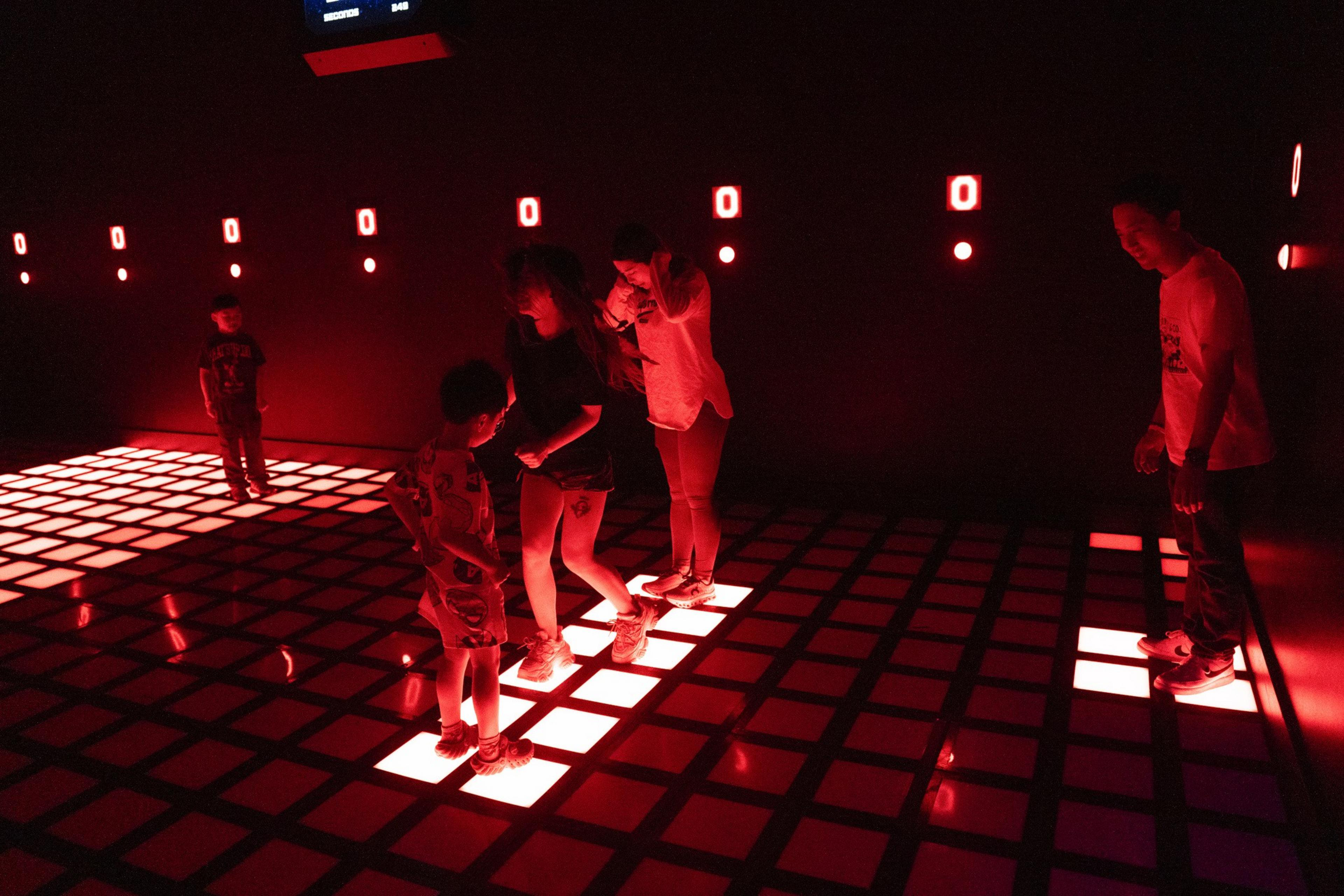 The image shows people standing on a glowing red grid floor in a dark room. The walls have small red lights and digital zeros displayed.