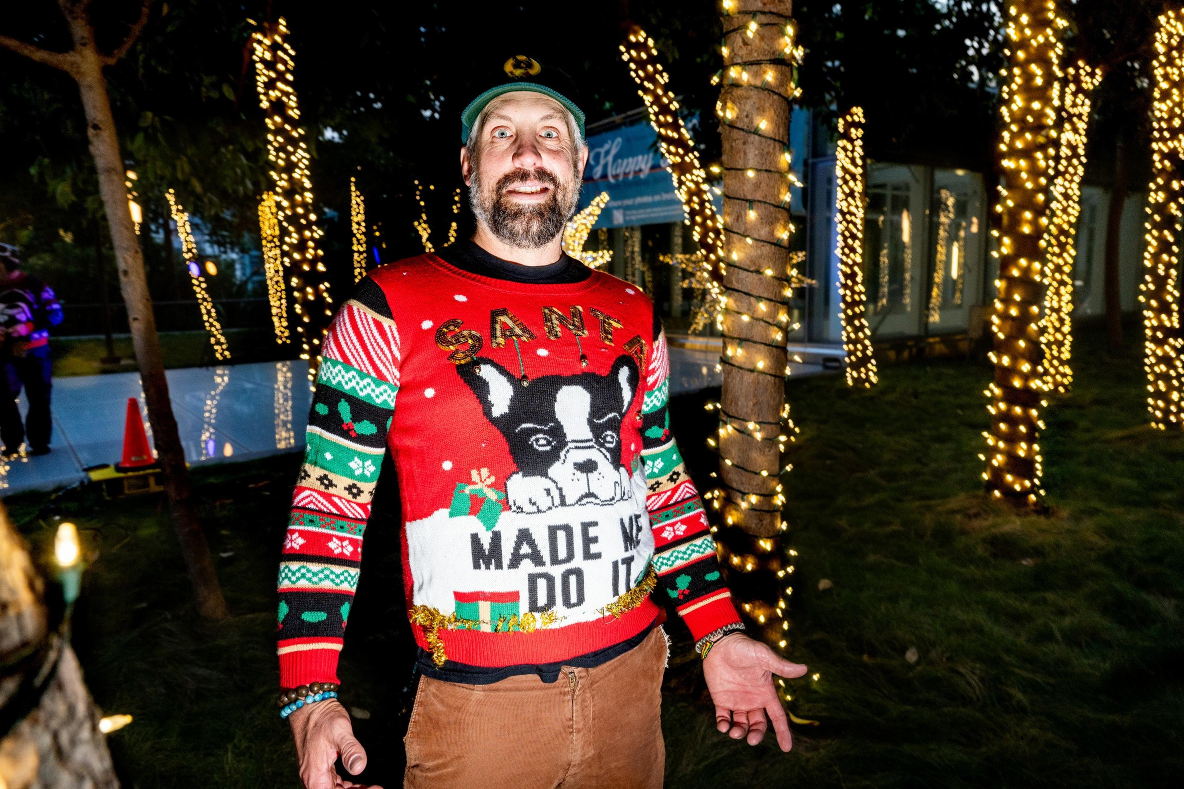 A man in a festive sweater with a dog, reading &quot;Santa made me do it,&quot; stands smiling among trees wrapped in white Christmas lights at night.