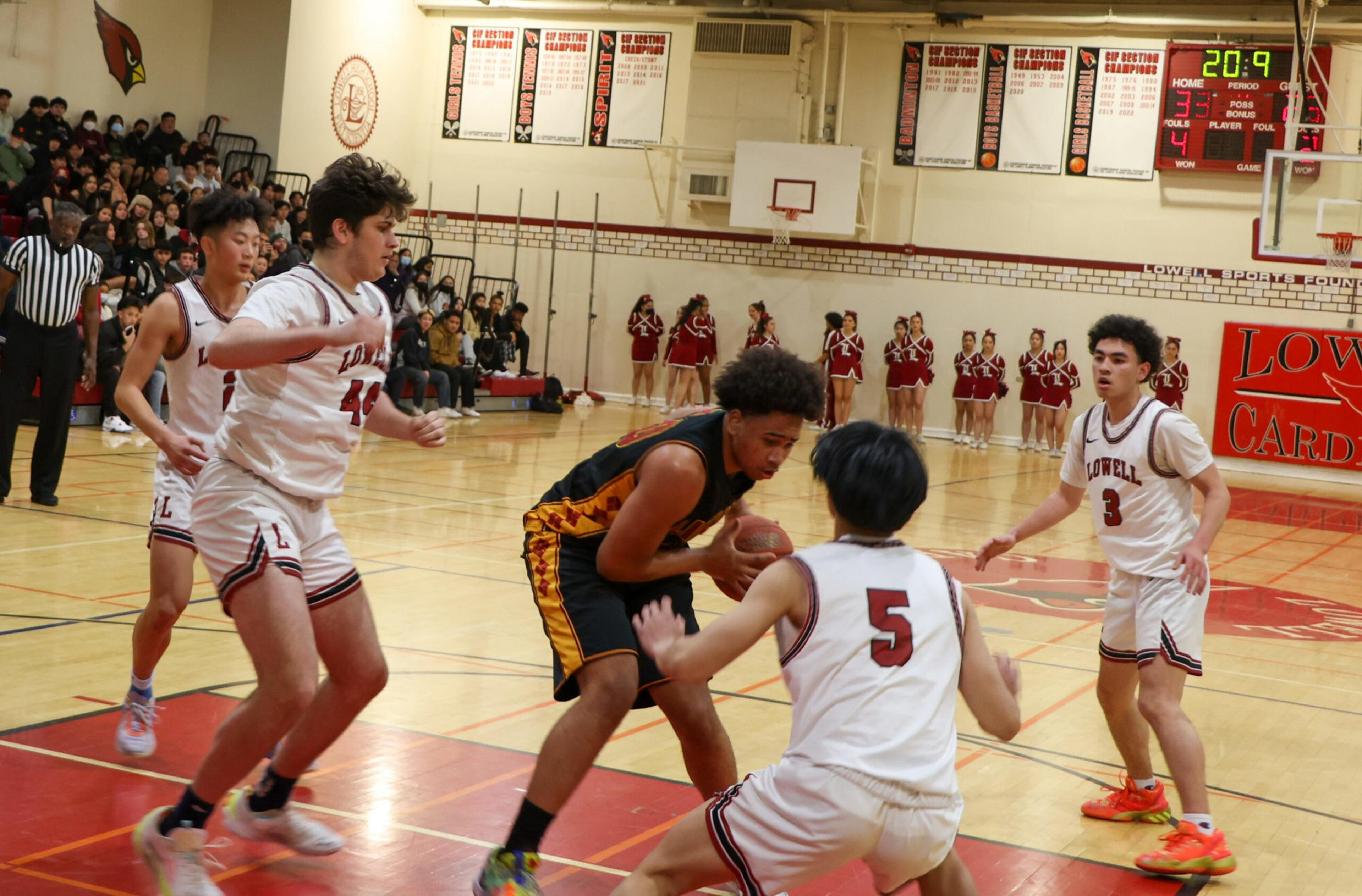 A basketball player in a black and orange uniform holds the ball while four players in white uniforms closely guard him on the court.