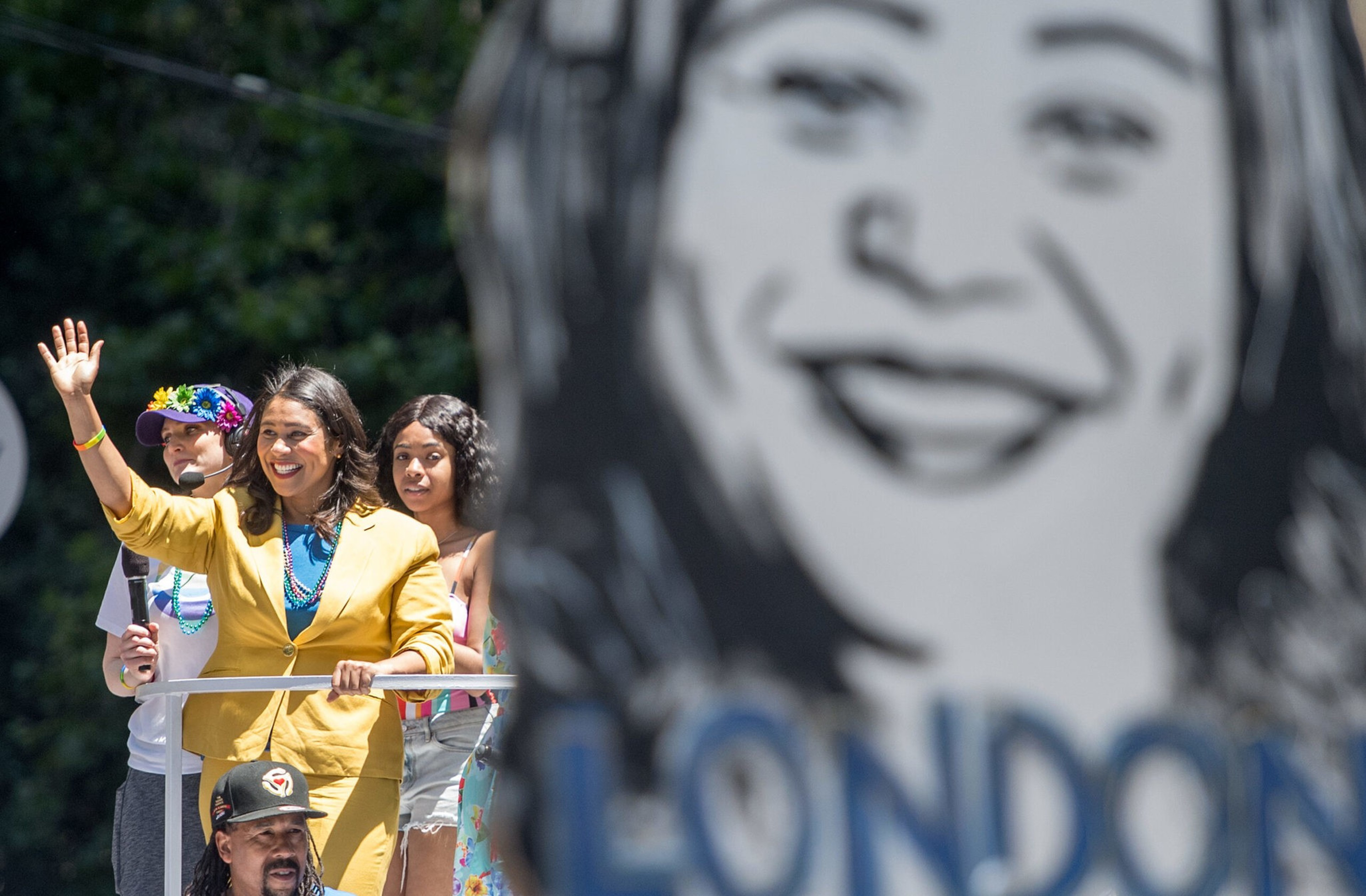A woman in a yellow suit waves and smiles, standing beside two younger people, with a large black-and-white face and "LONDON" blurred in the foreground.