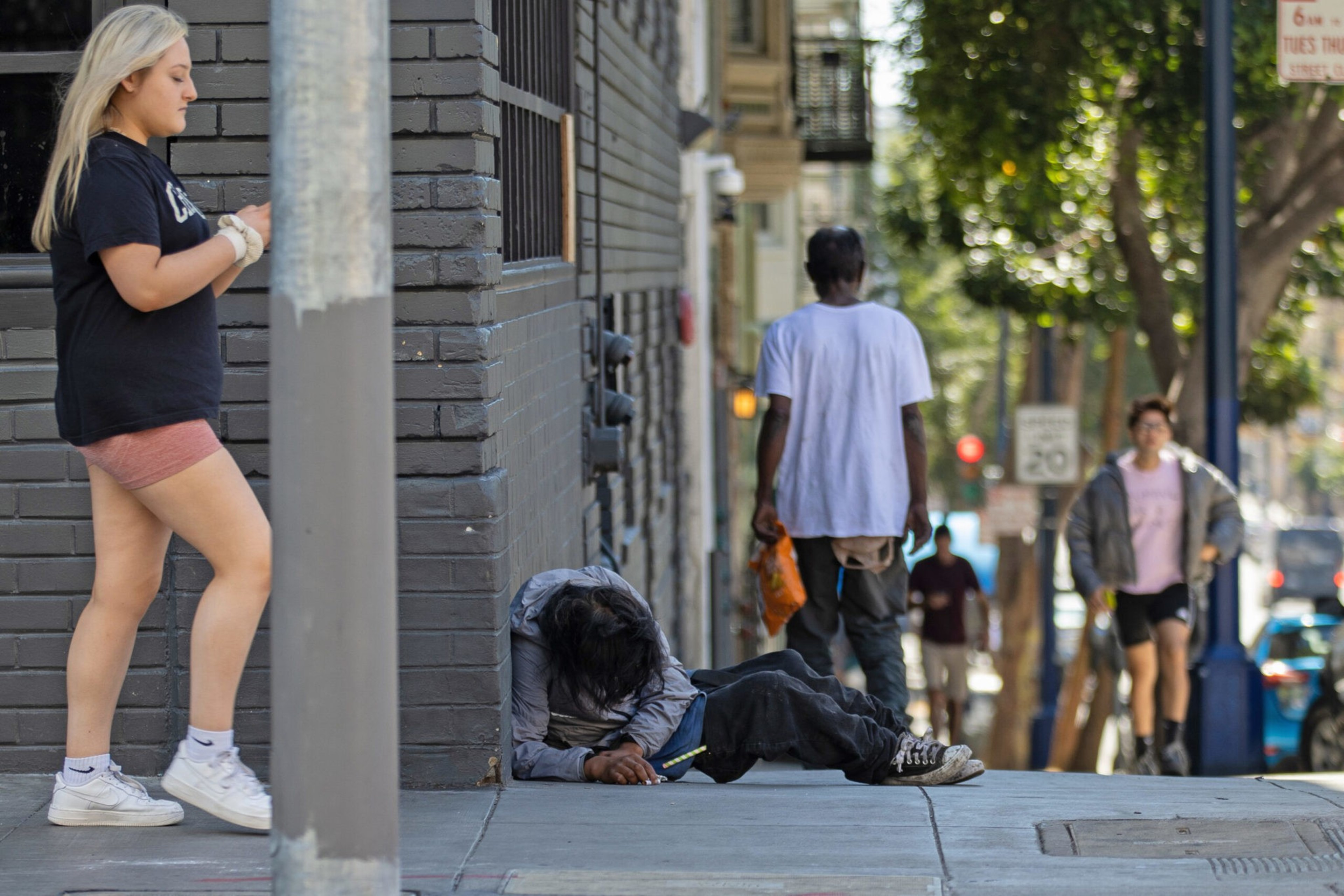 A person lies on the sidewalk against a brick wall, while others walk past on the urban street nearby.