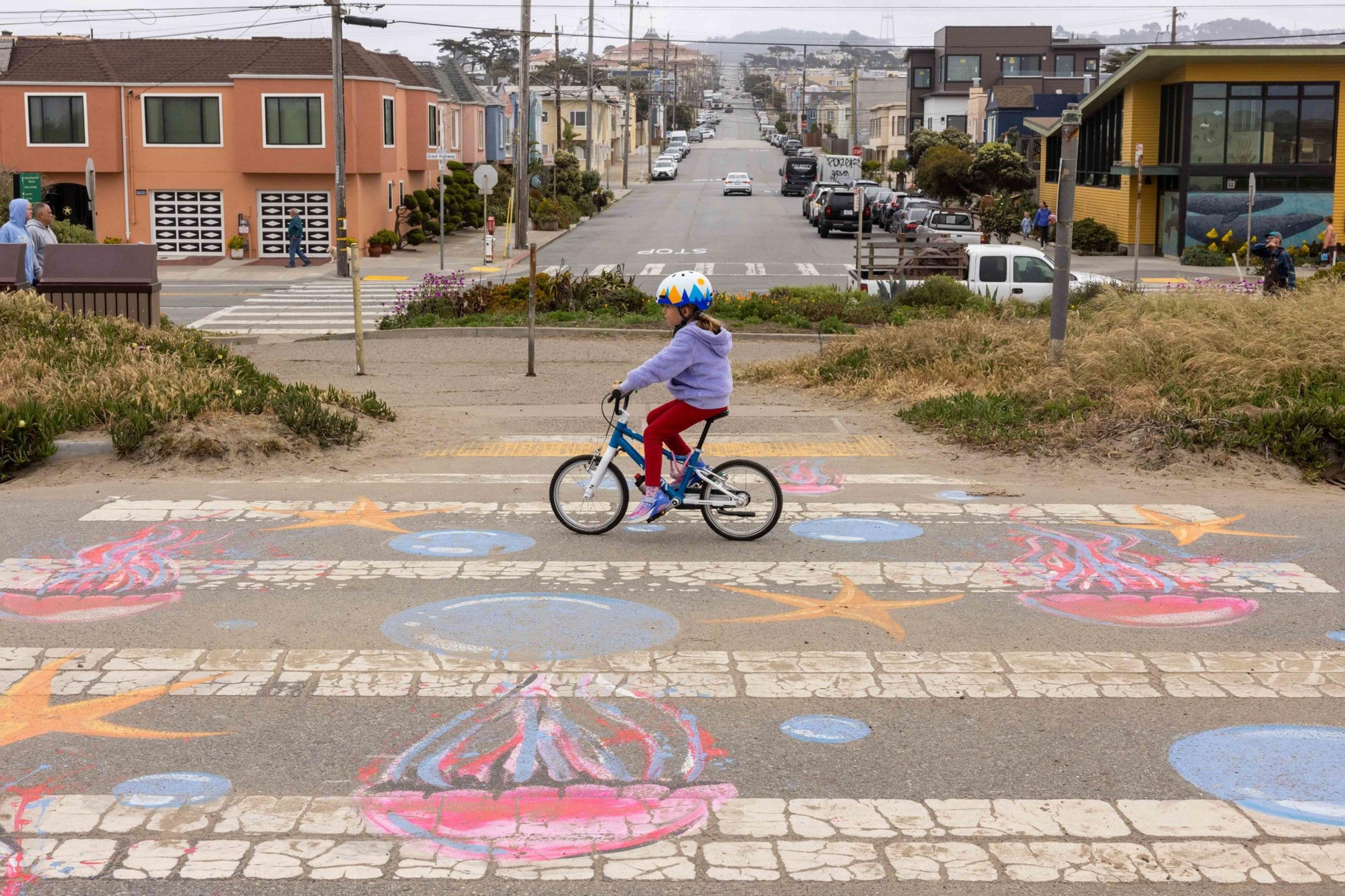 A child wearing a colorful helmet rides a bike across a crosswalk decorated with bright chalk drawings of jellyfish and bubbles, set in a suburban street.