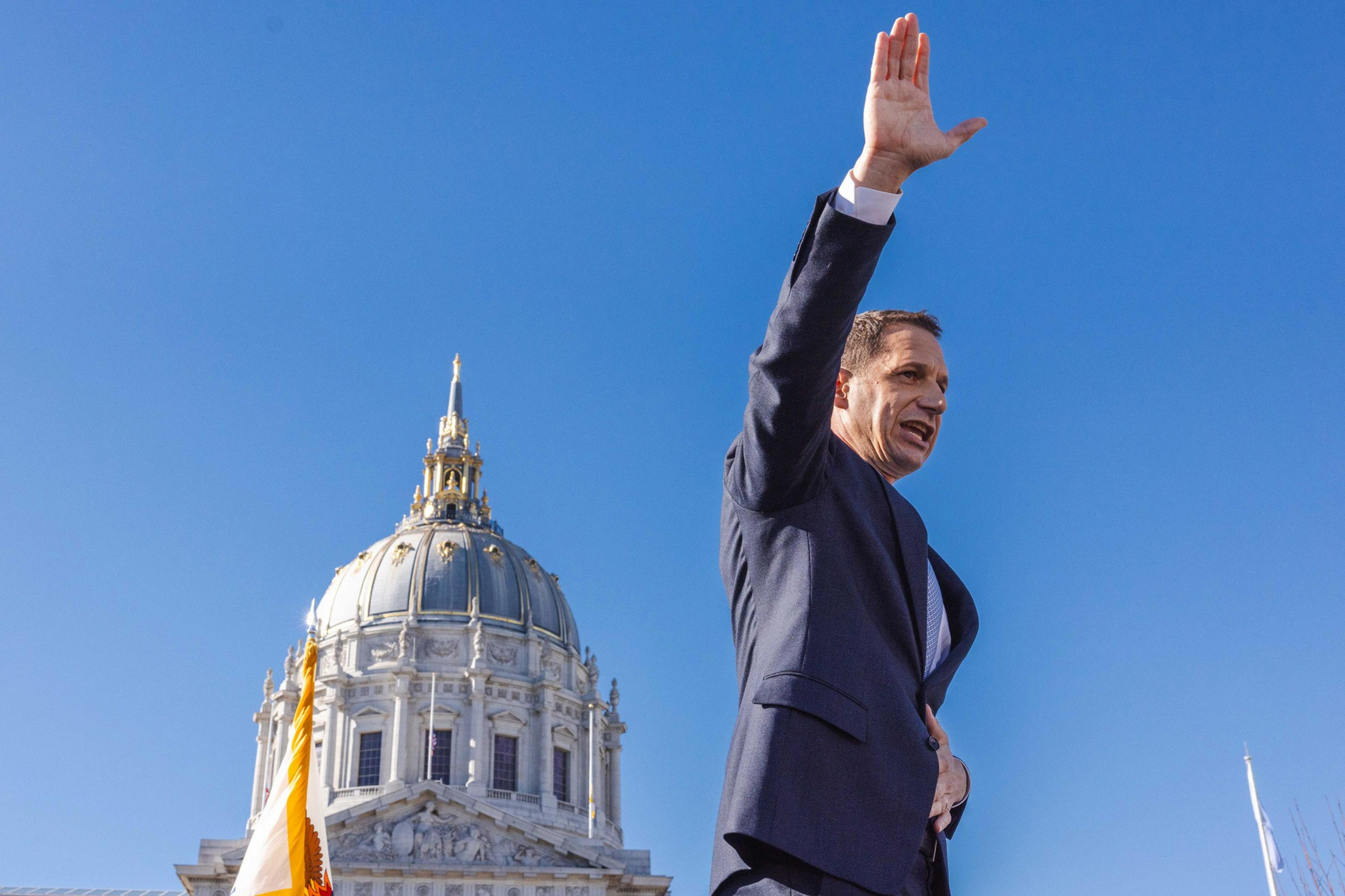A person in a suit stands against a clear blue sky, raising one hand. Behind them is an ornate, domed building.