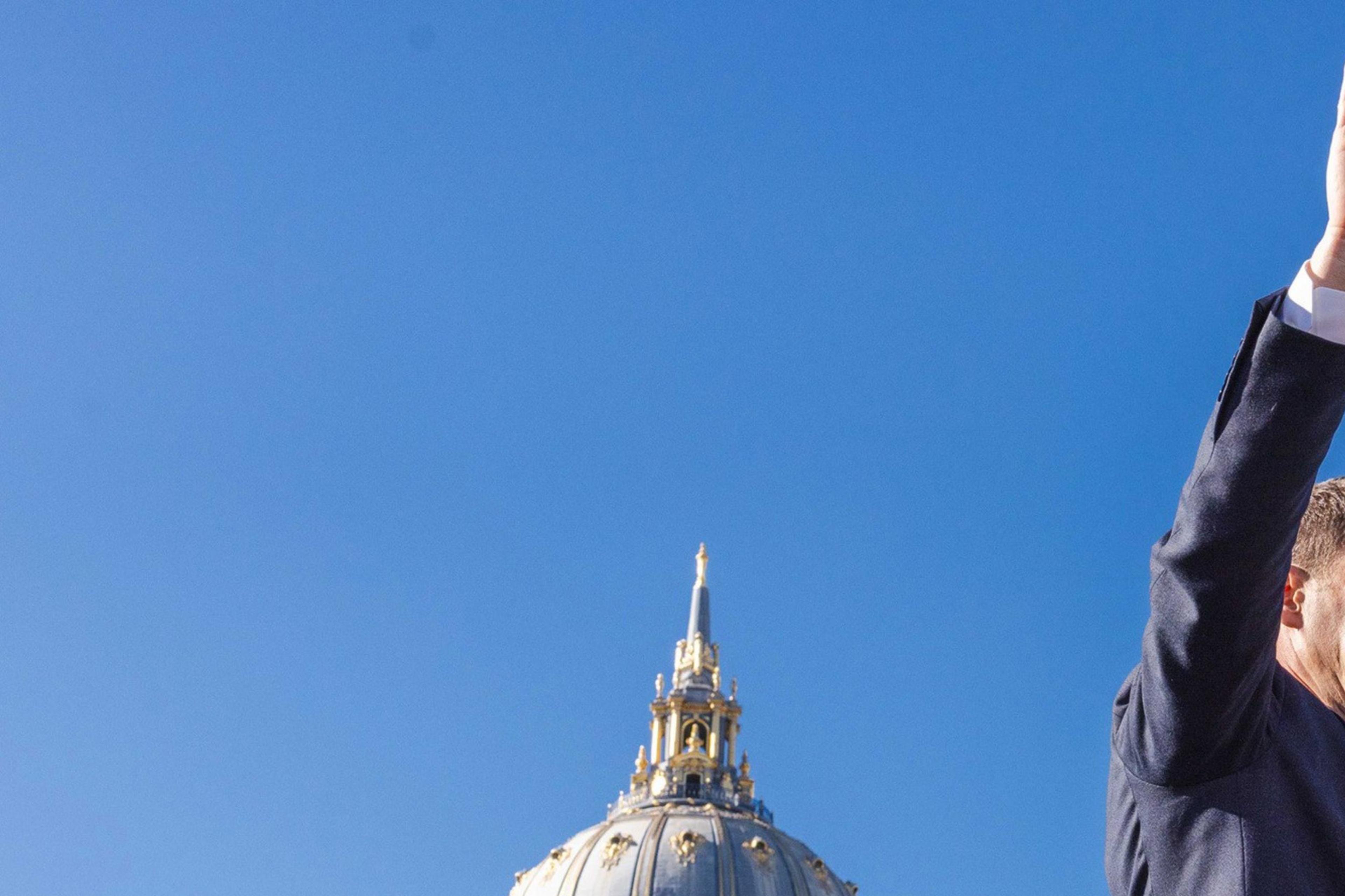 A person in a suit stands against a clear blue sky, raising one hand. Behind them is an ornate, domed building.
