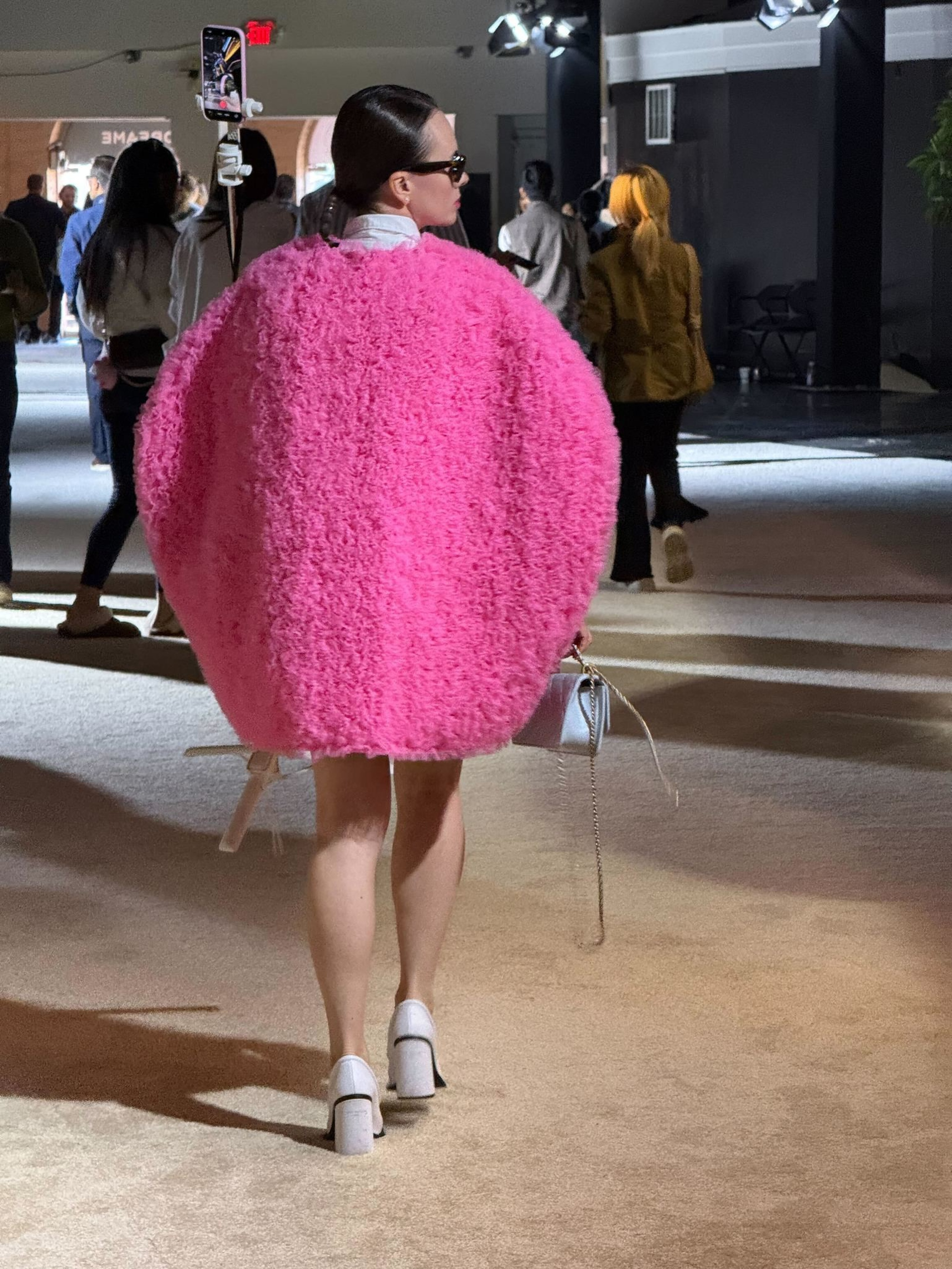 A person walks wearing a large, round, bright pink fuzzy coat, white heels, and sunglasses, holding a small bag in a well-lit indoor space.