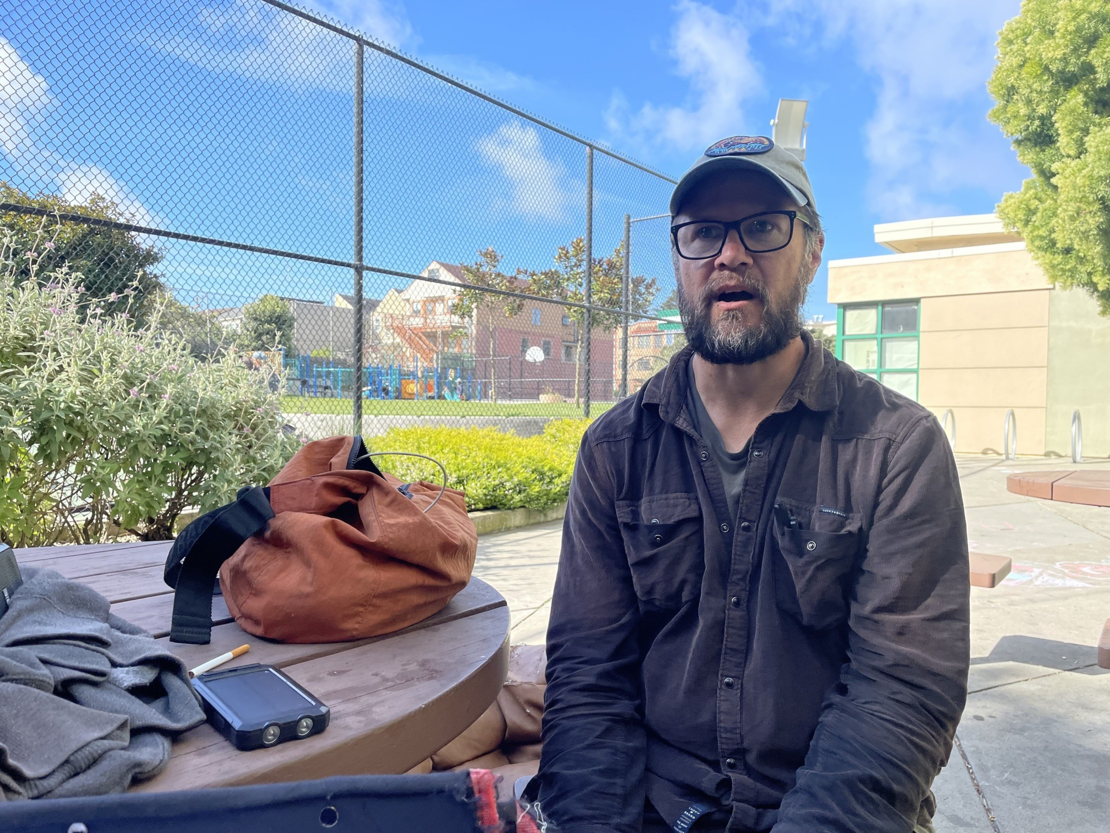 A man sits at a table in a park with belongings in a wagon nearby.