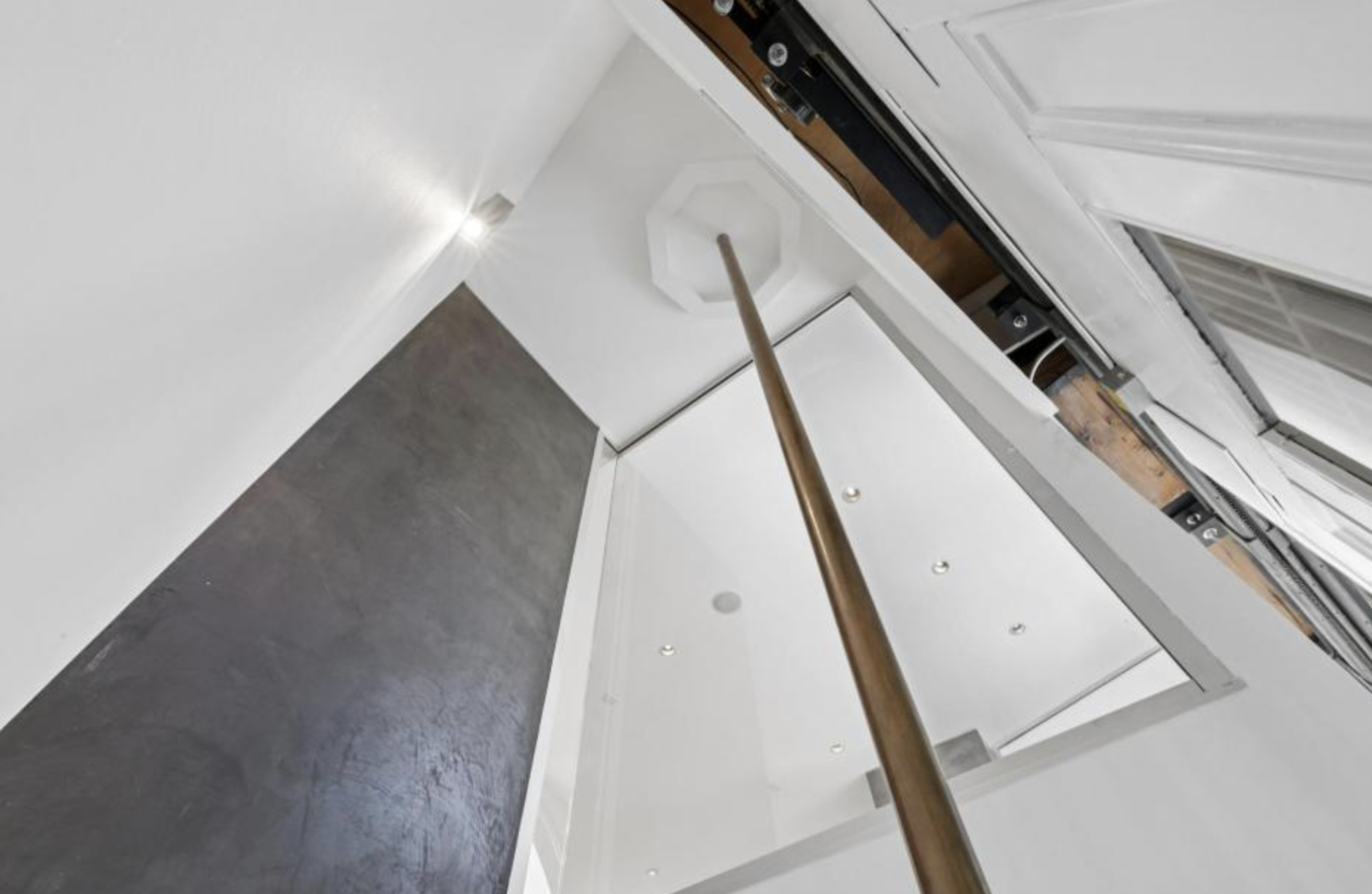 A tall, narrow space with white walls and ceiling, a black textured wall on one side, and a bronze-colored vertical pole reaching up into a small octagonal loft opening.