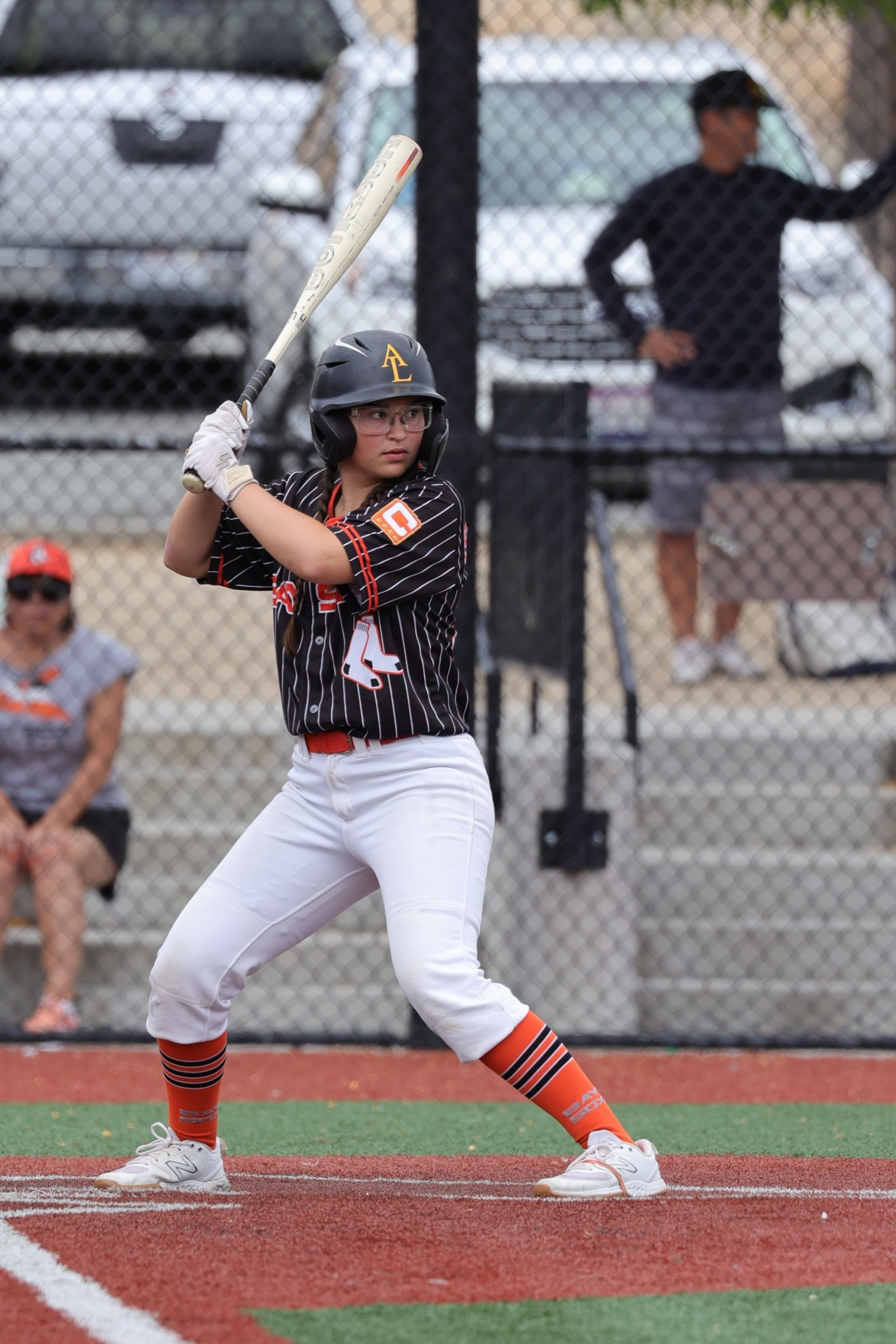 A young baseball player in a black and orange uniform stands poised to bat on a red and green field with spectators behind a chain-link fence.