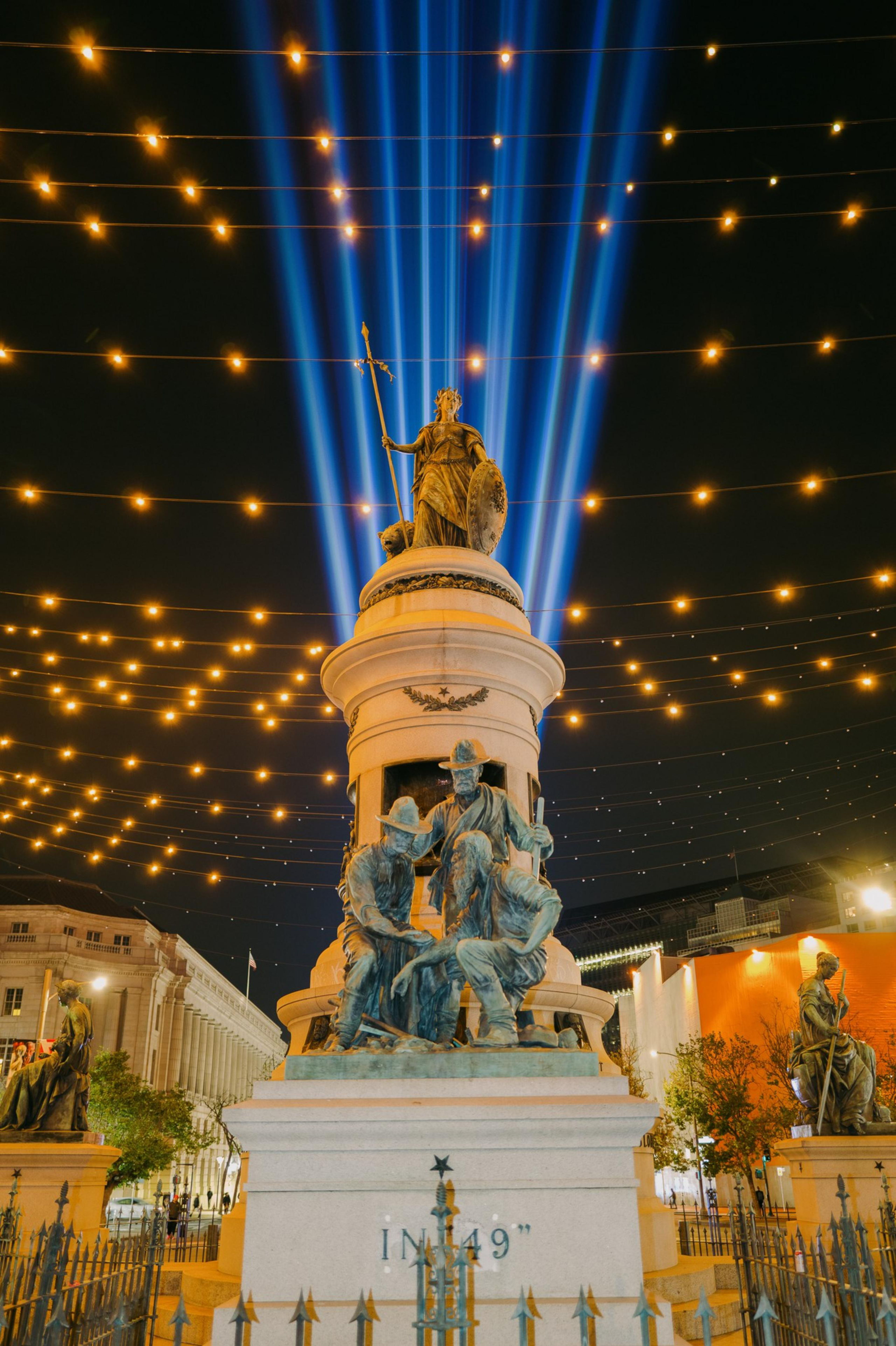 A tall monument with statues is surrounded by blue vertical light beams and string lights glowing against the night sky.