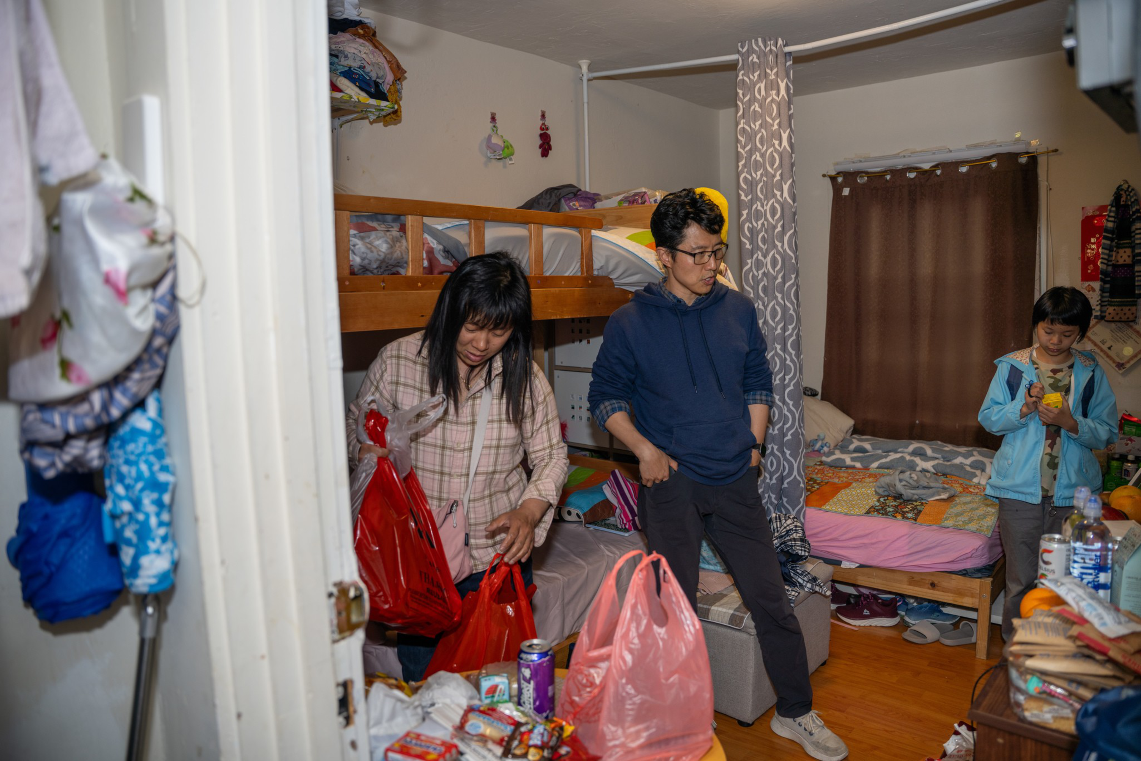 Three people are in a cluttered bedroom with bunk beds, unpacking bags filled with food and household items.