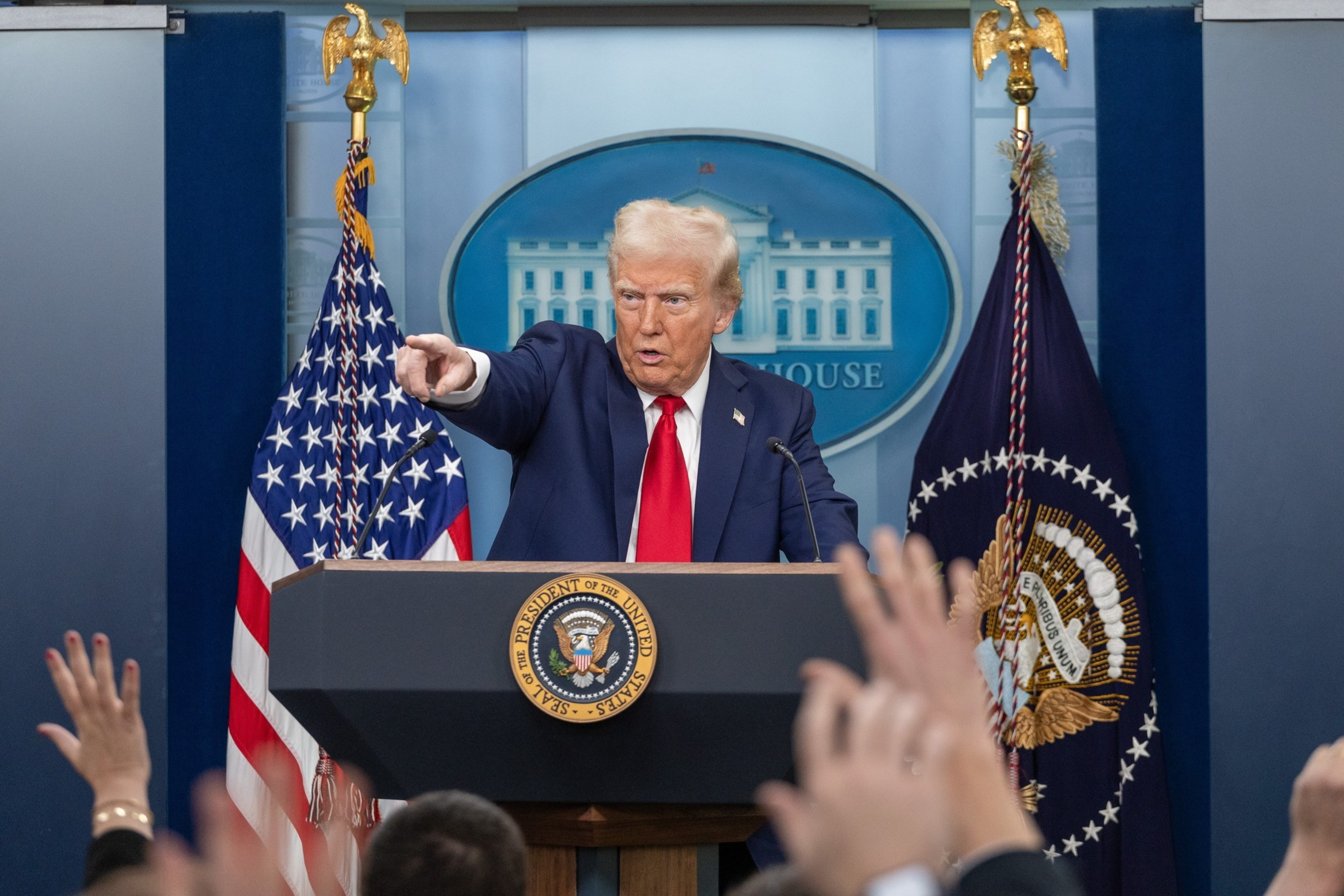 A man in a suit and red tie points forward while standing at a podium with the presidential seal, with raised hands visible in the audience.