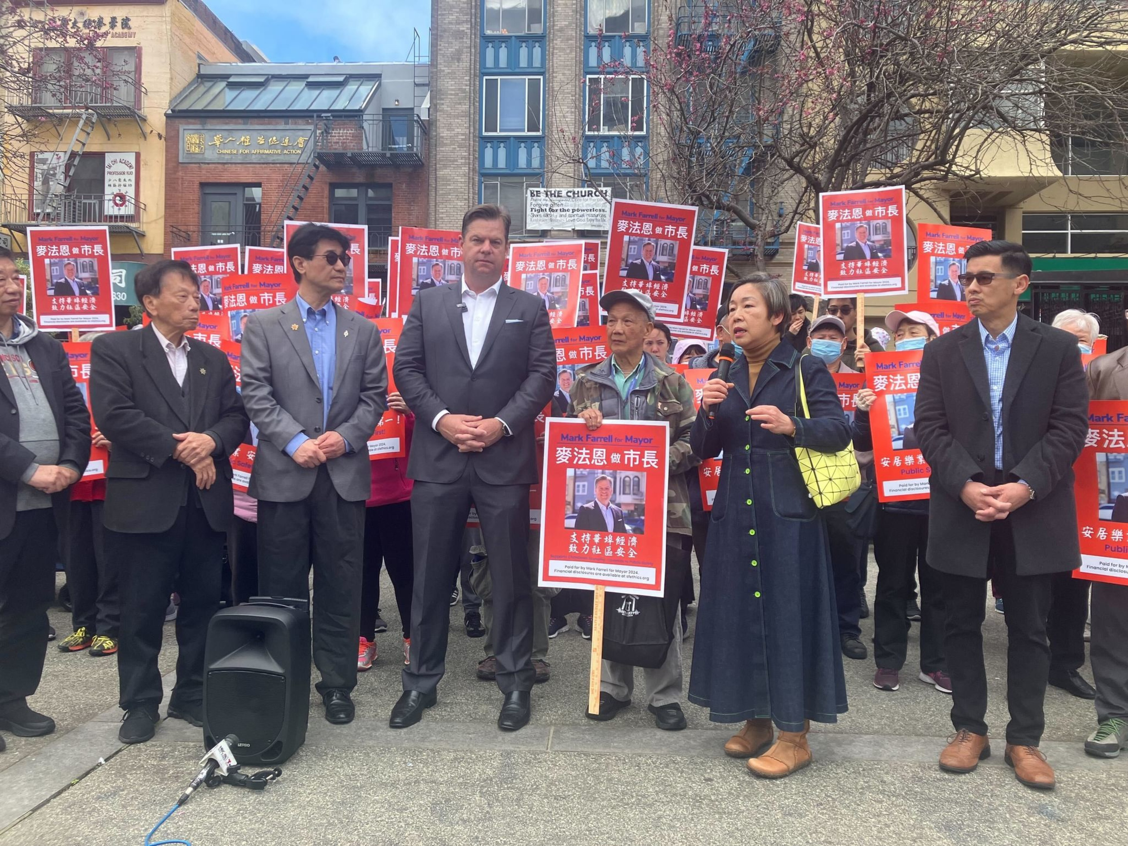 A group stands outdoors holding red signs with a man's photo and Chinese text, with a speaker addressing them.