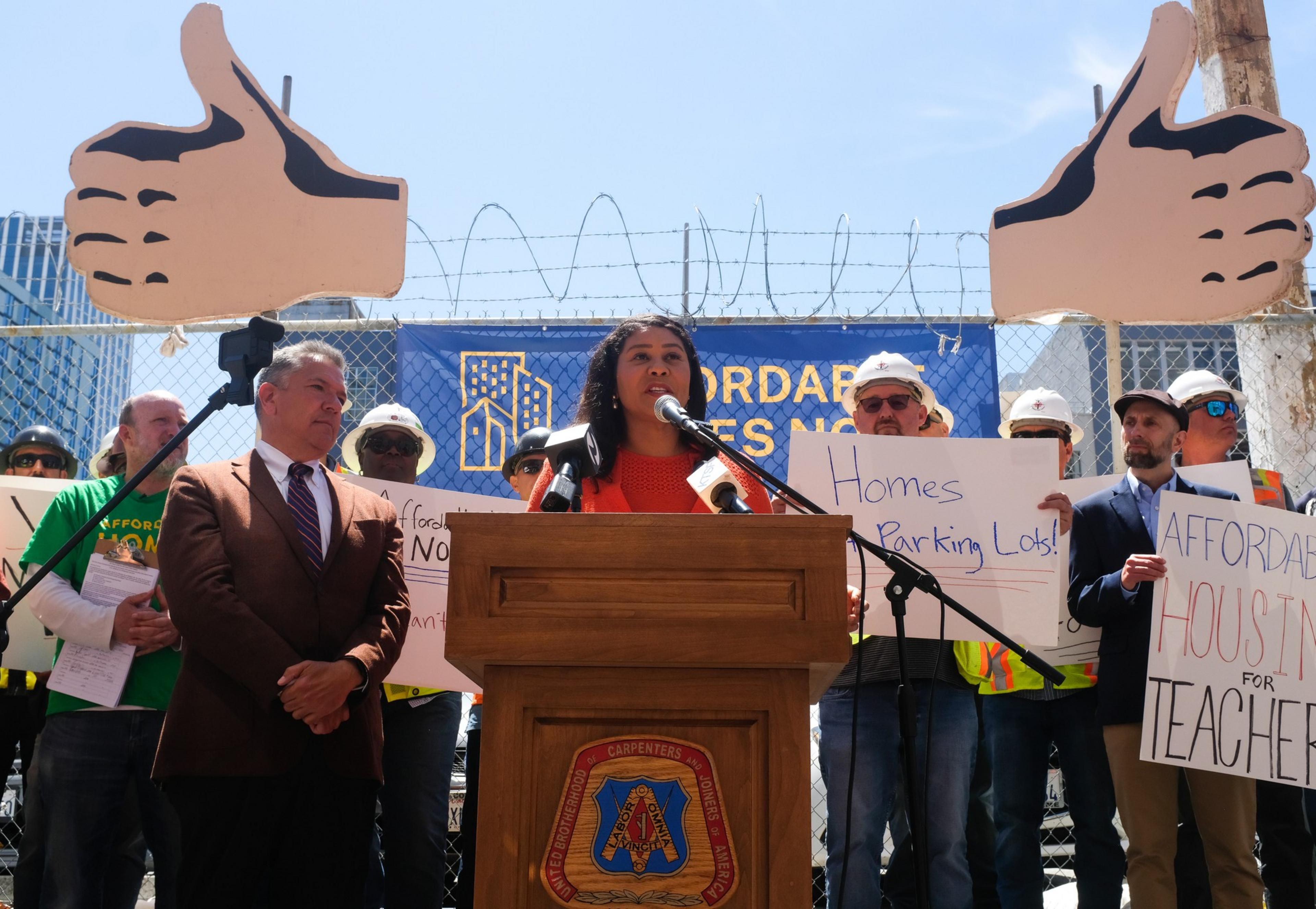 A woman speaks at a podium surrounded by people holding signs about affordable housing. Two large foam thumbs-up signs are in the background.