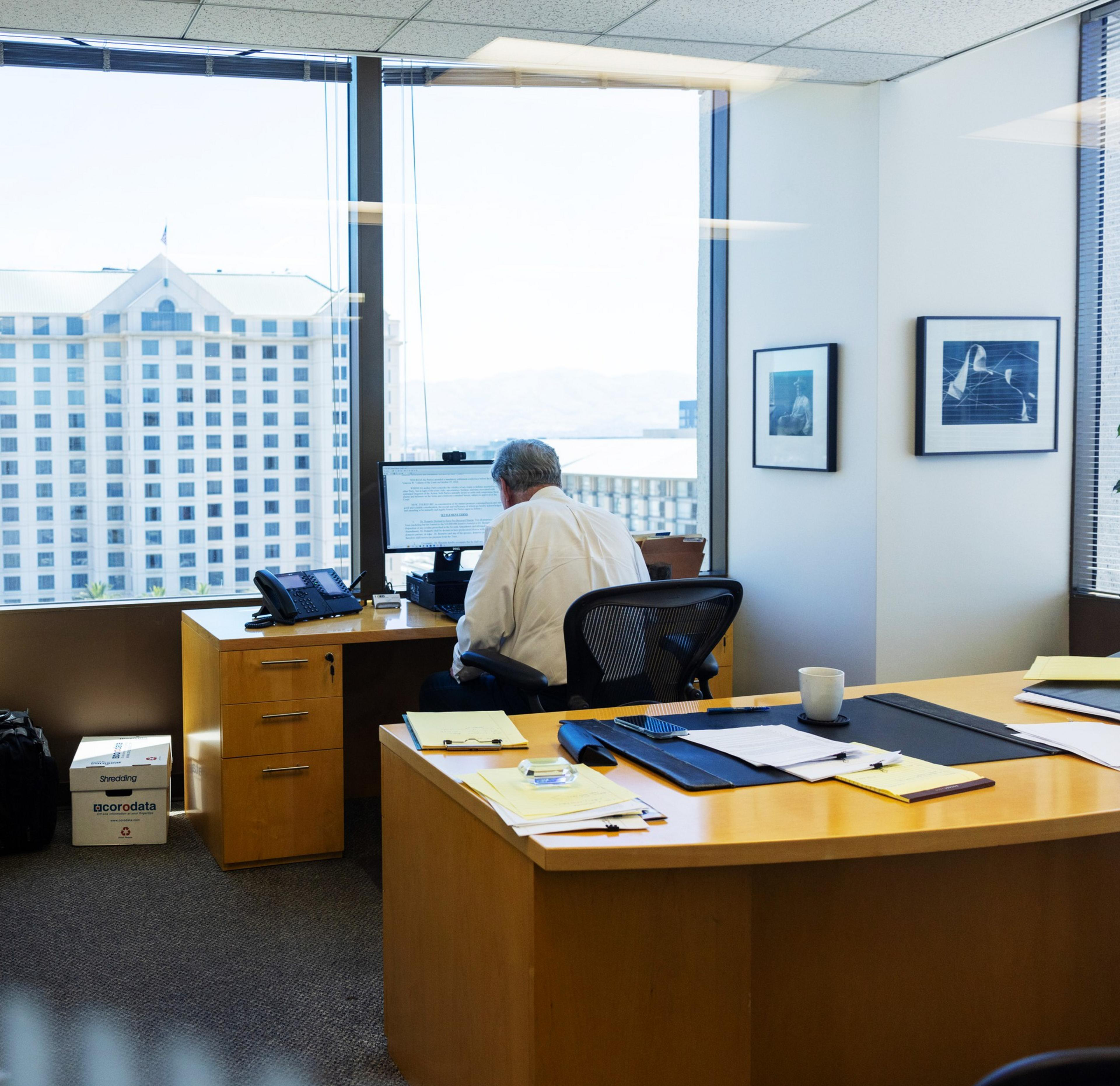 An office with a man in a white shirt working at a computer by a large window overlooking a building. The desk is cluttered with papers, folders, and a coffee mug.