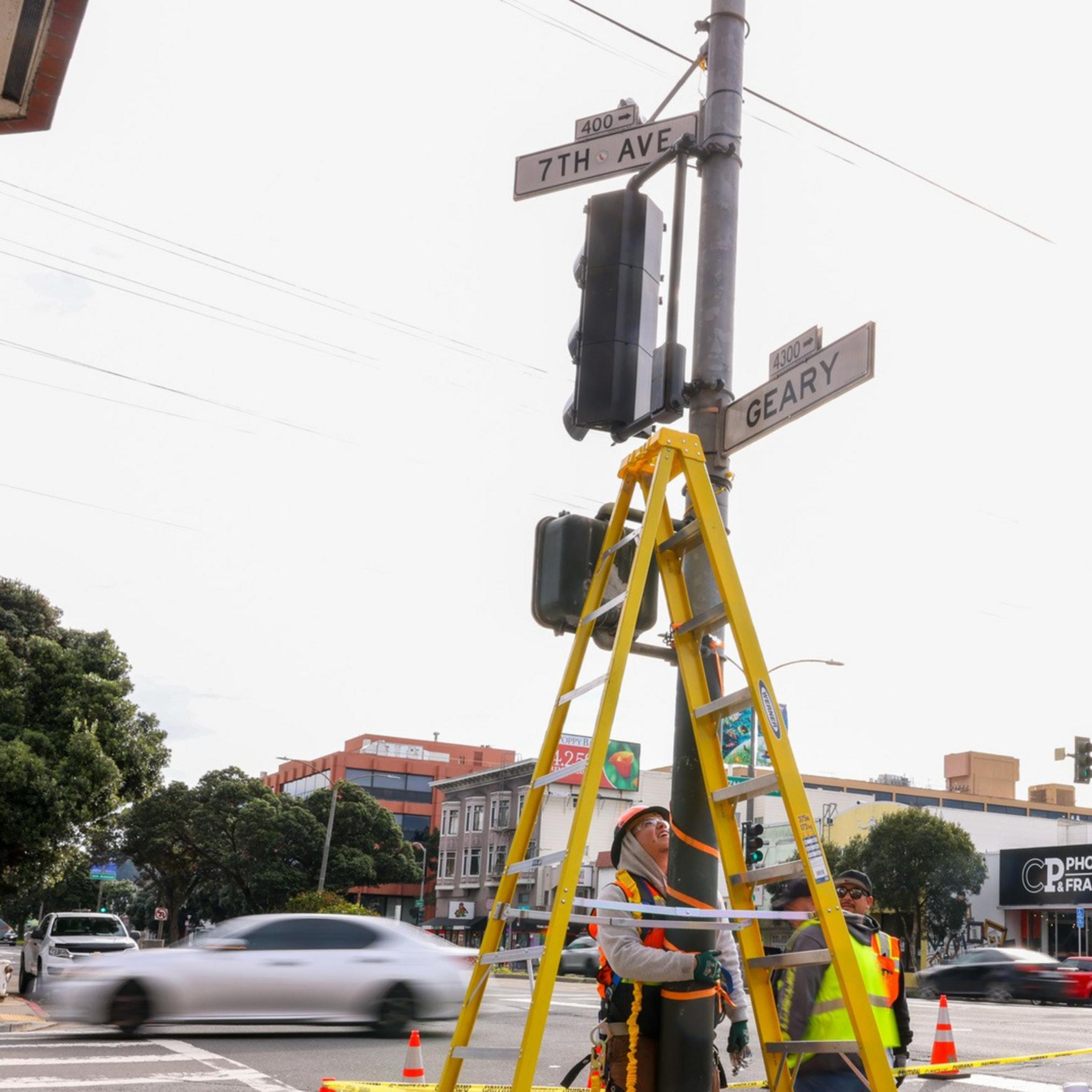 Construction workers in safety gear are standing at a traffic intersection with a ladder propped against a pole, surrounded by cones and caution tape.