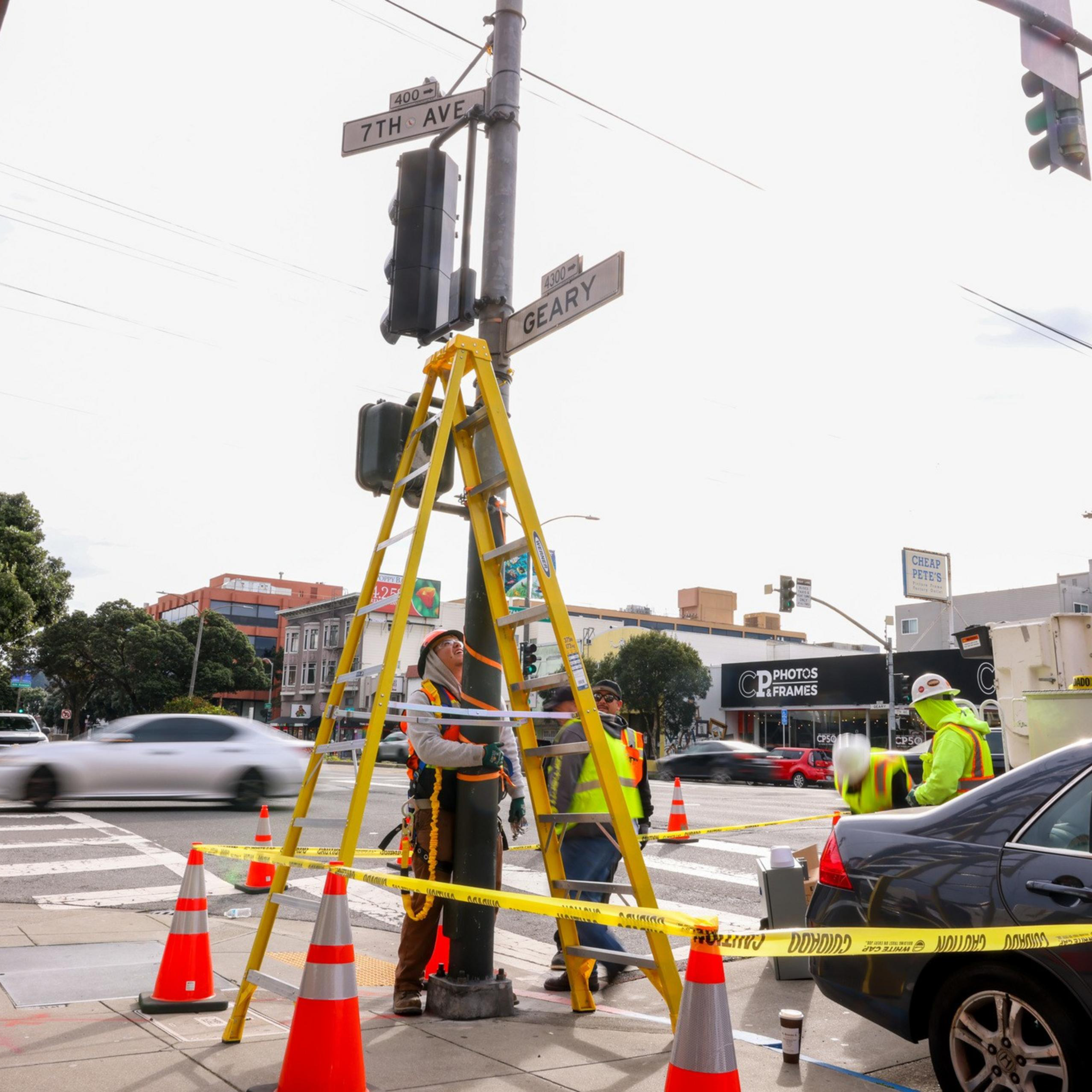 Construction workers in safety gear are standing at a traffic intersection with a ladder propped against a pole, surrounded by cones and caution tape.