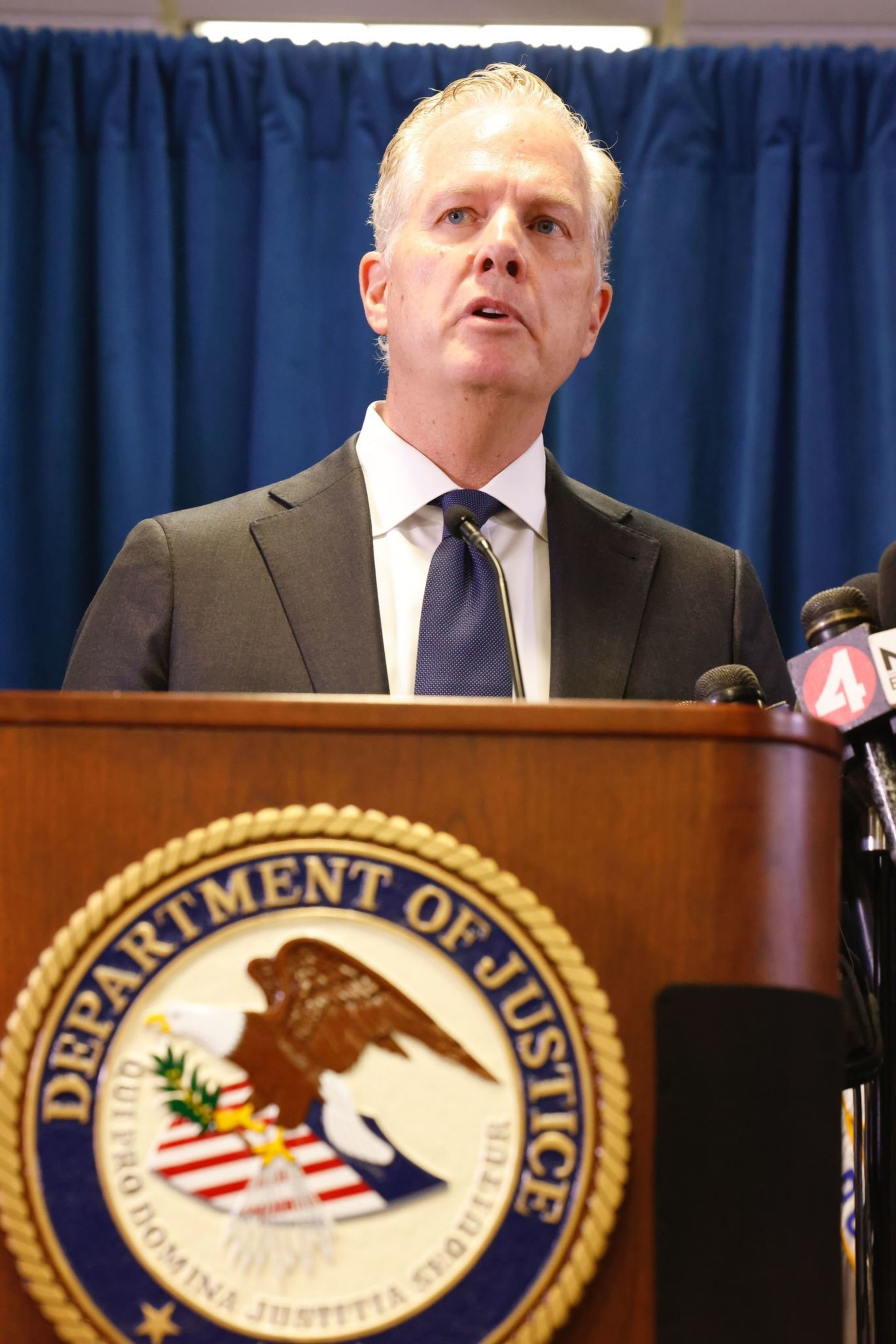 A man in a suit speaks at a podium with the Department of Justice emblem. There's a blue curtain background and several microphones in view.