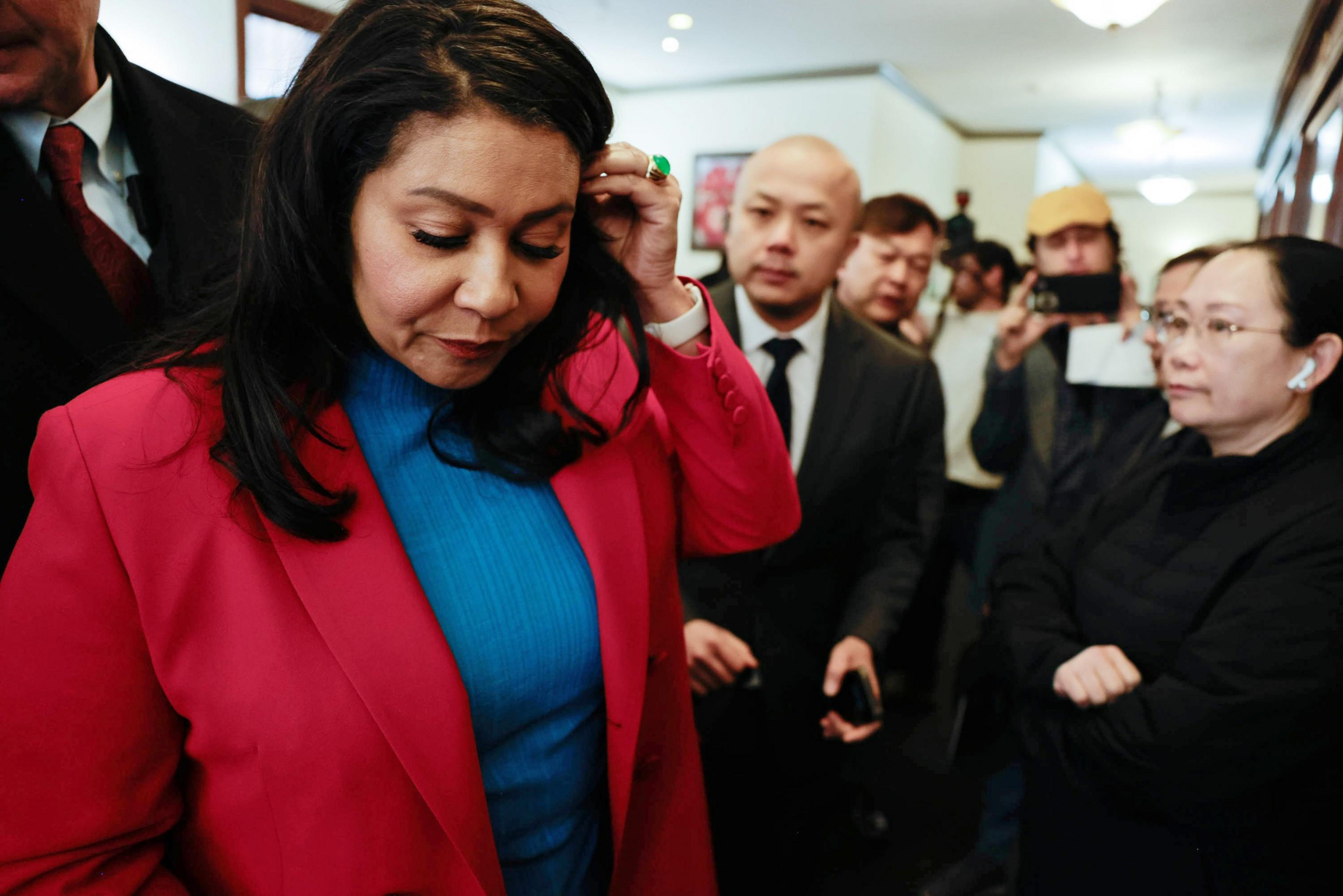 A woman in a red blazer is walking with her head down, surrounded by people and photographers.